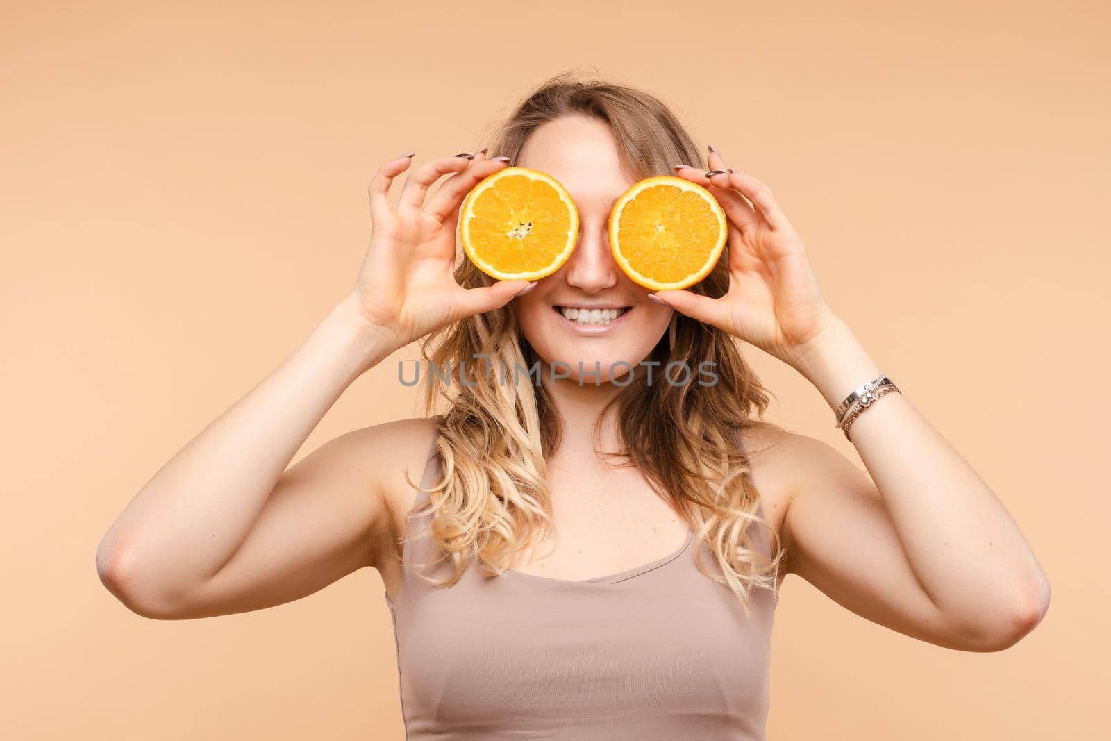 Front view of cheerful young blonde posing with fresh oranges on isolated background. Funny girl keeping fruit and closing eyes in studio. Concept of happiness and health.