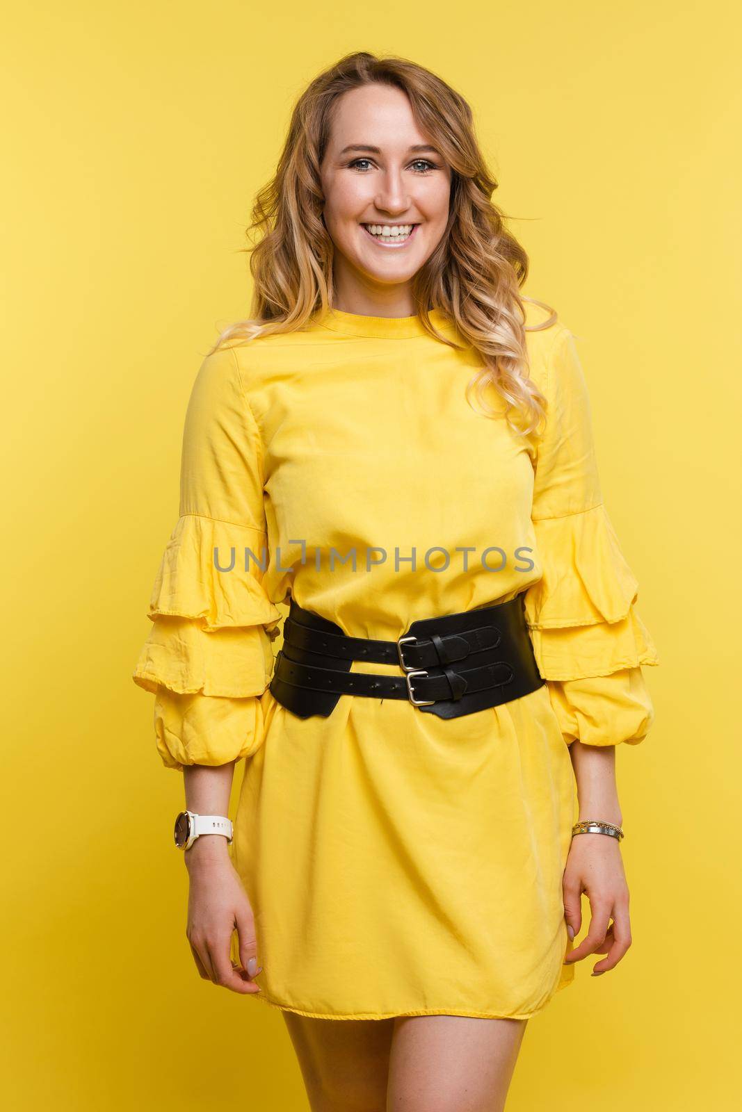 Studio portrait of fashionable brunette lady in white dress with flowers and beige heels posing with bent leg. Smiling at camera. holding skirt. Isolate on yellow background.