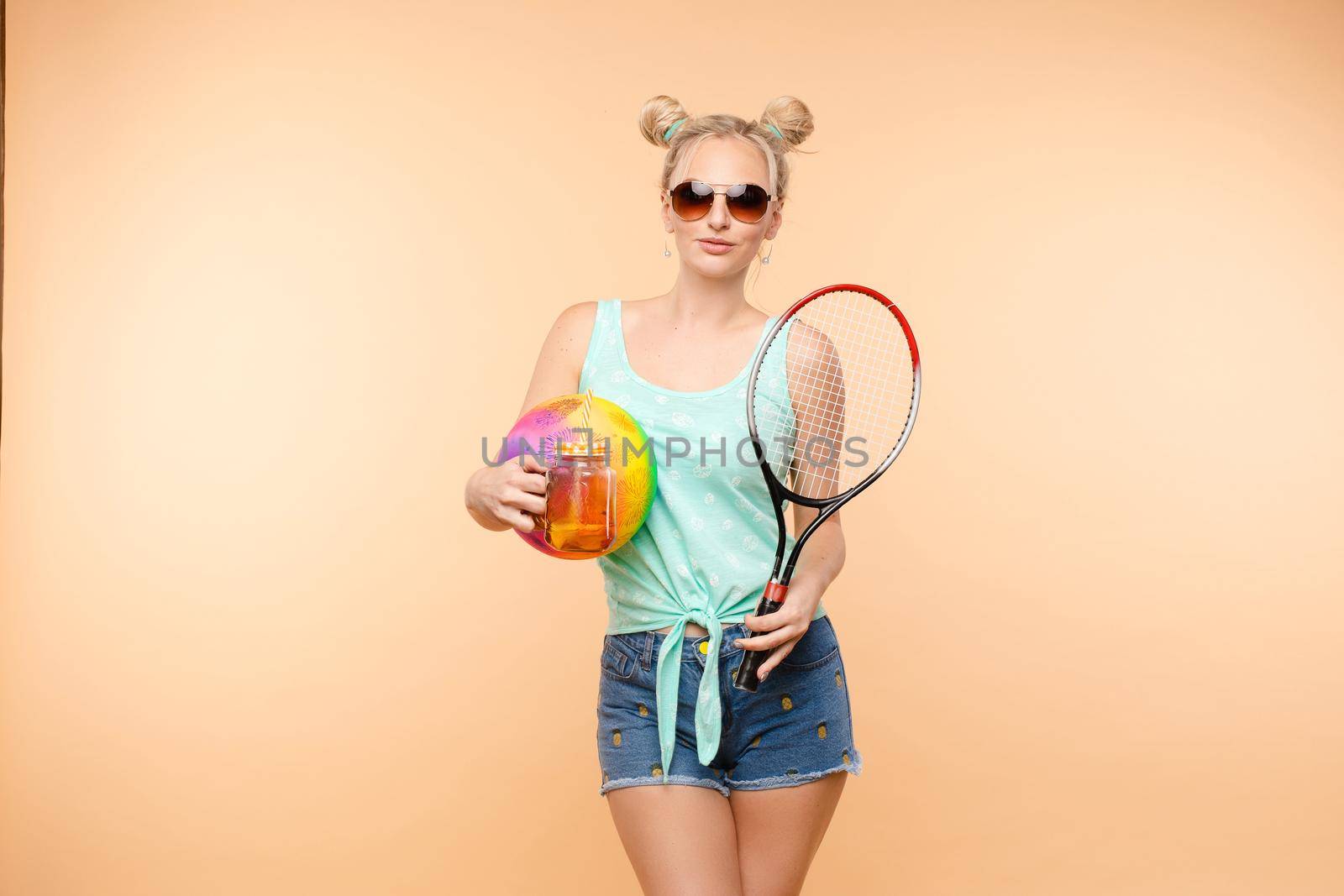 Side view of sporty blonde drinking fresh juice, doing sport and posing in studio. Young model keeping ball and racket and smiling on isolated background. Concept of energy and power.