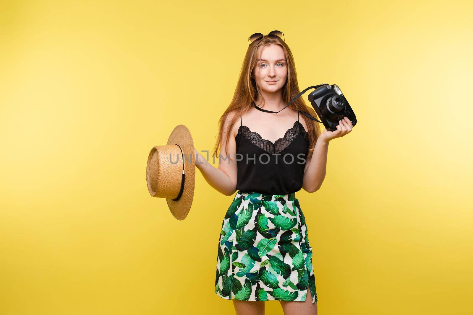 Smiling elegance travel young woman posing with camera and straw hat medium long shot. Beautiful tourist female ready to summer trip isolated at yellow studio background