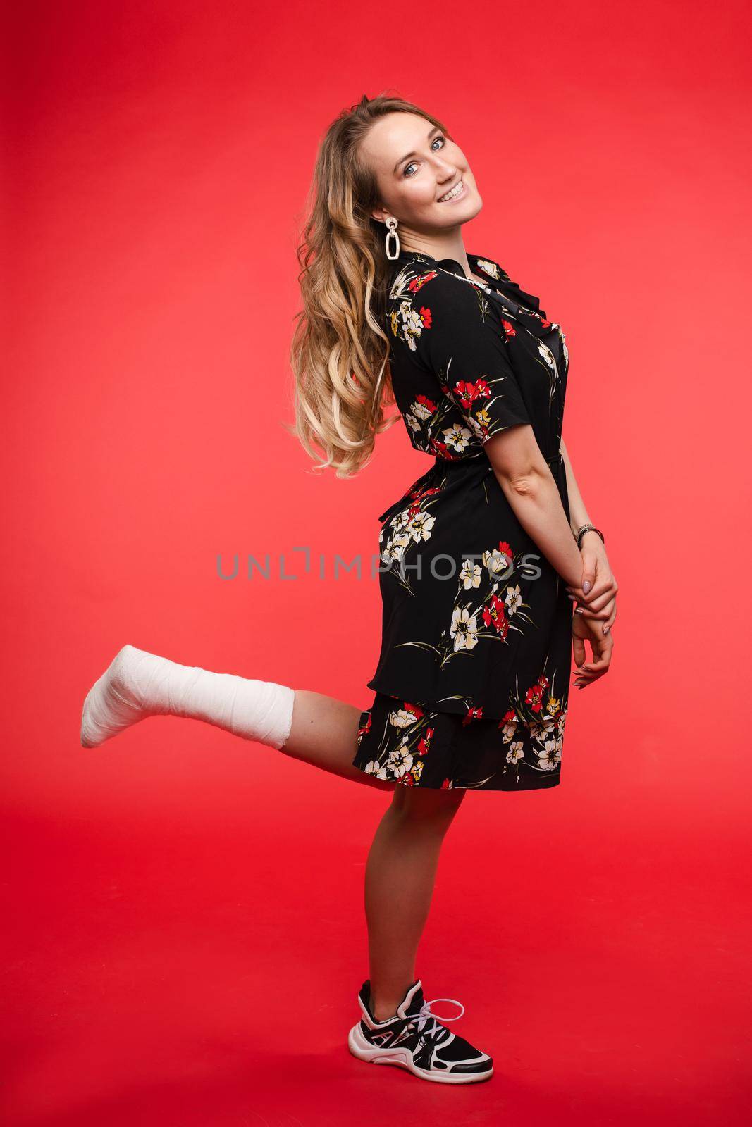 Full length studio shot of beautiful young woman with wavy hair wearing elegant flowered dress and sneaker with broken right left leg in plaster. Isolate on red background.