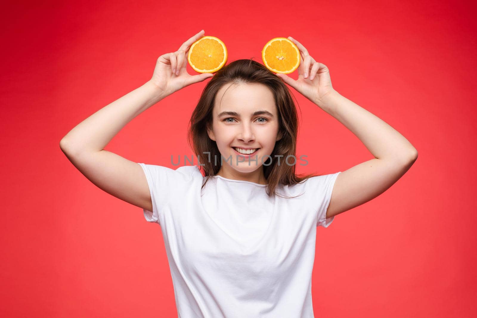 Studio headshot of young funny brunette with hairstyle and red lips holding halved oranges on eyes against bright yellow background.