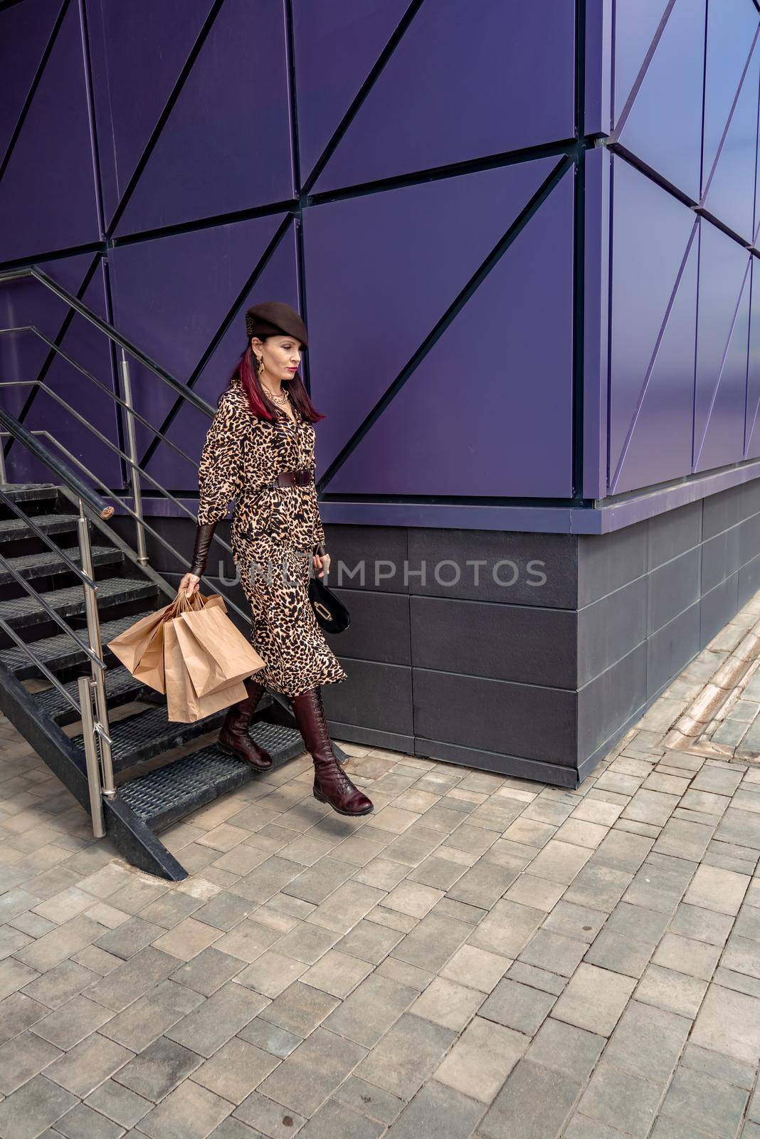 A happy shopaholic girl keeps her bags near the shopping center. A woman near the store is happy with her purchases, holding bags. Dressed in a leopard print dress. Consumer concept. by Matiunina