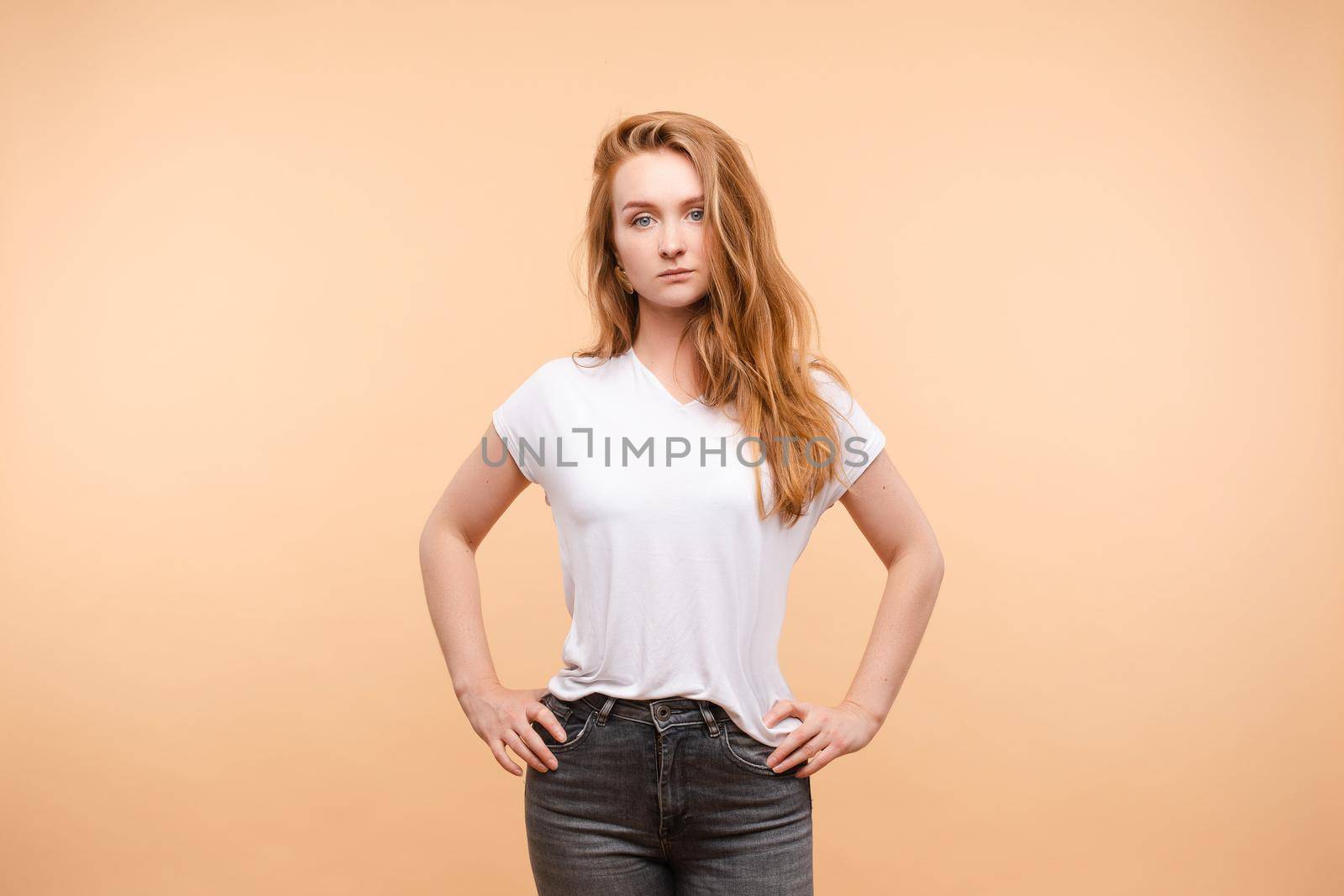 Front view of young serious woman wearing white shirt and jeans standing on isolated background. Calm confident female looking at camera in studio. Concept of casual outfit and expression.