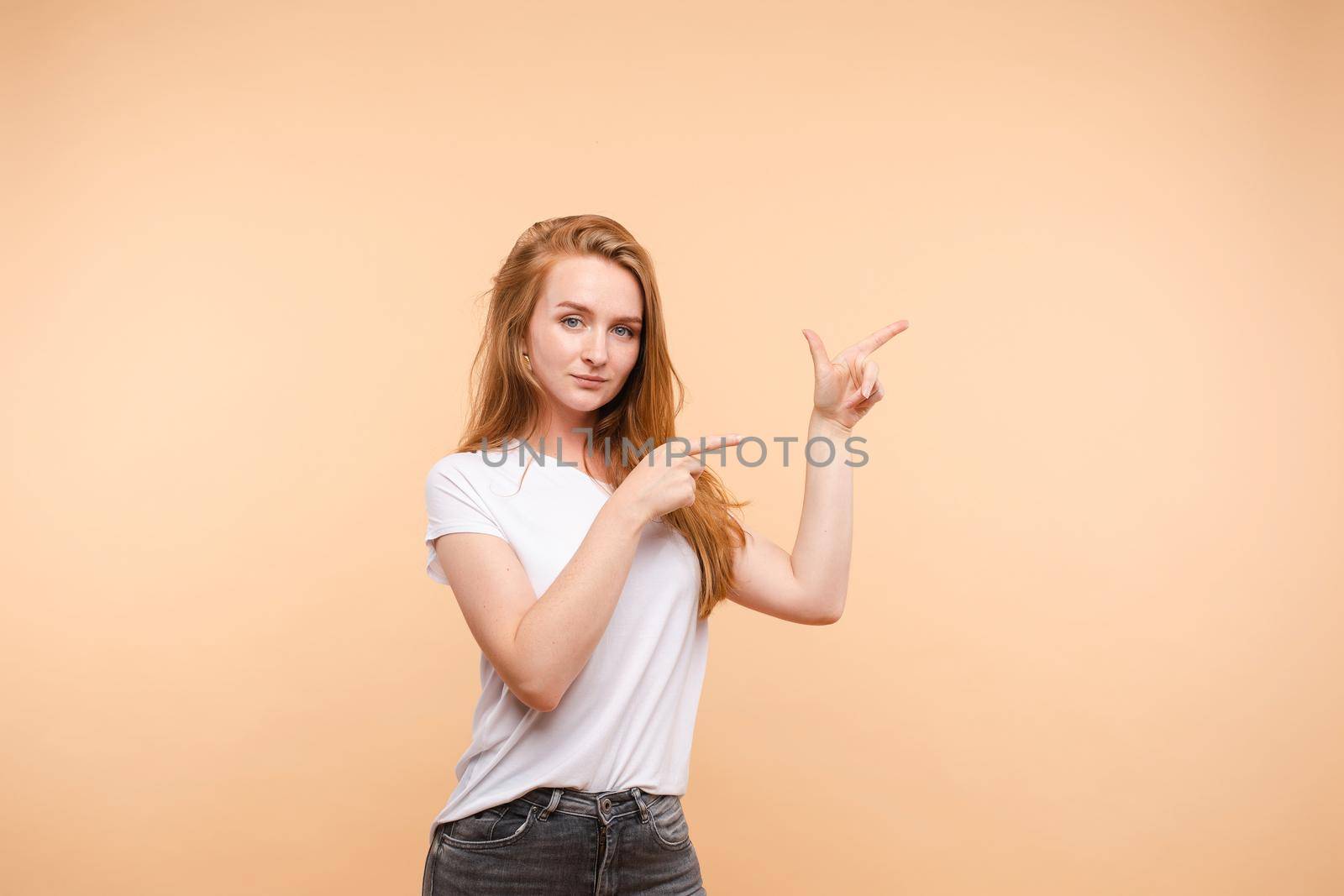 Studio portrait of ginger young woman in plain white t-shirt and jeans demonstrating something in the air indicating blank space with her fingers on both arms. She is looking at camera. Isolate.