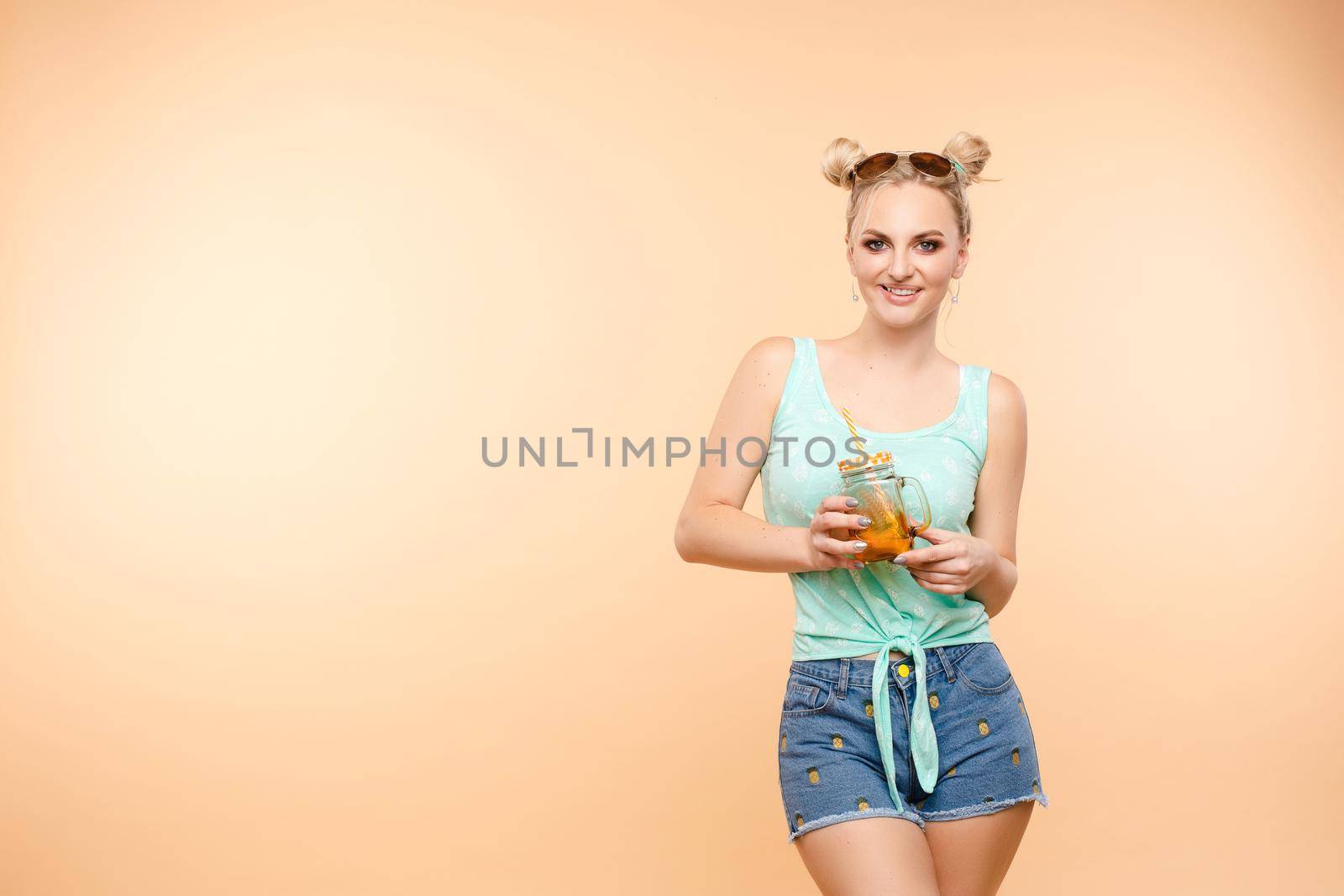 Young happy lady standing on one leg and holding other one up. Blond girl in casual clothes enjoying tasty fruit cocktail. Beautiful woman in sunglasses holding jar with citrus lemonade in hands.