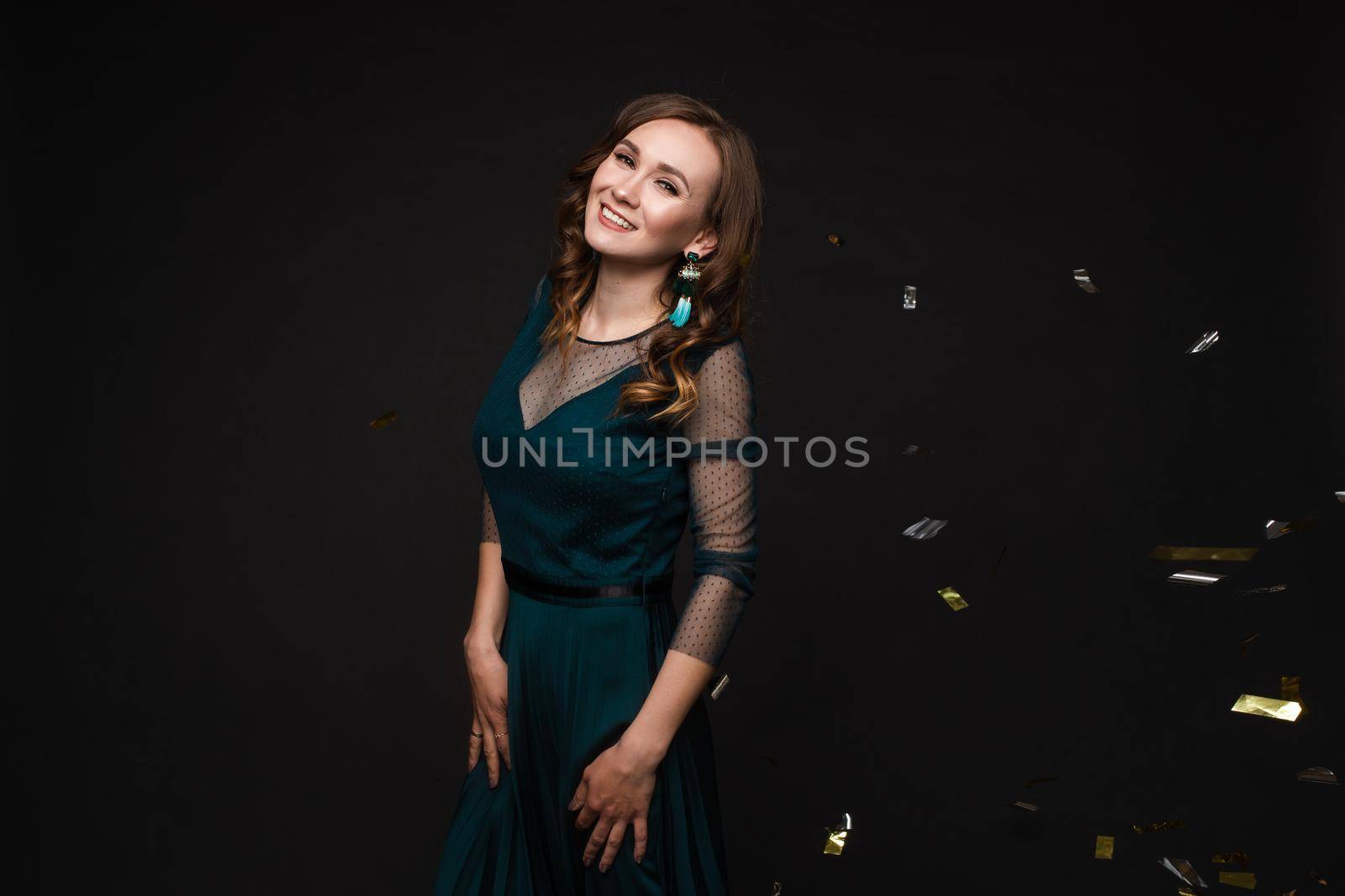 Happy young girl looking surprisingly while golden confetti falling. Beautiful woman in emerald elegant long dress smiling and dancing among decorations. Gorgeous lady celebrating birthday on party.