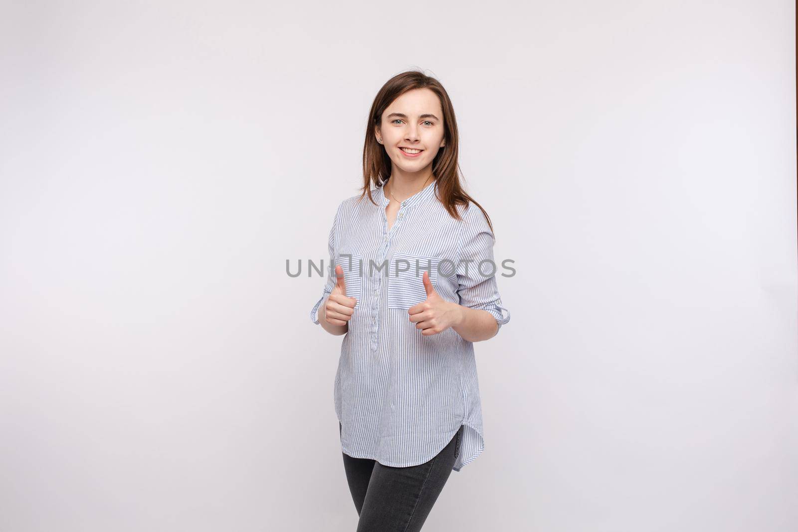 Studio portrait of pretty brunette woman in shirt showing thumbs up and smiling at camera with pleasure and satisfaction. Isolate on white.