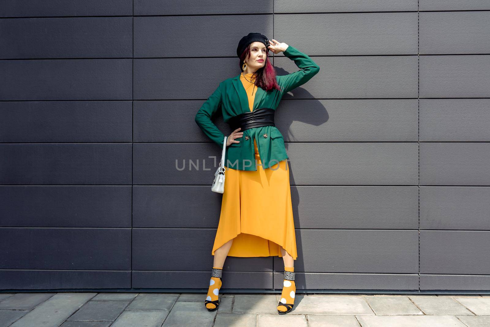 Young woman in bright clothes, yellow skirt and green jacket. Yellow socks in sandals, beret on the head, hair with the color of magenta. Caucasian female fashion model standing against gray wall background, open with copy space. by Matiunina