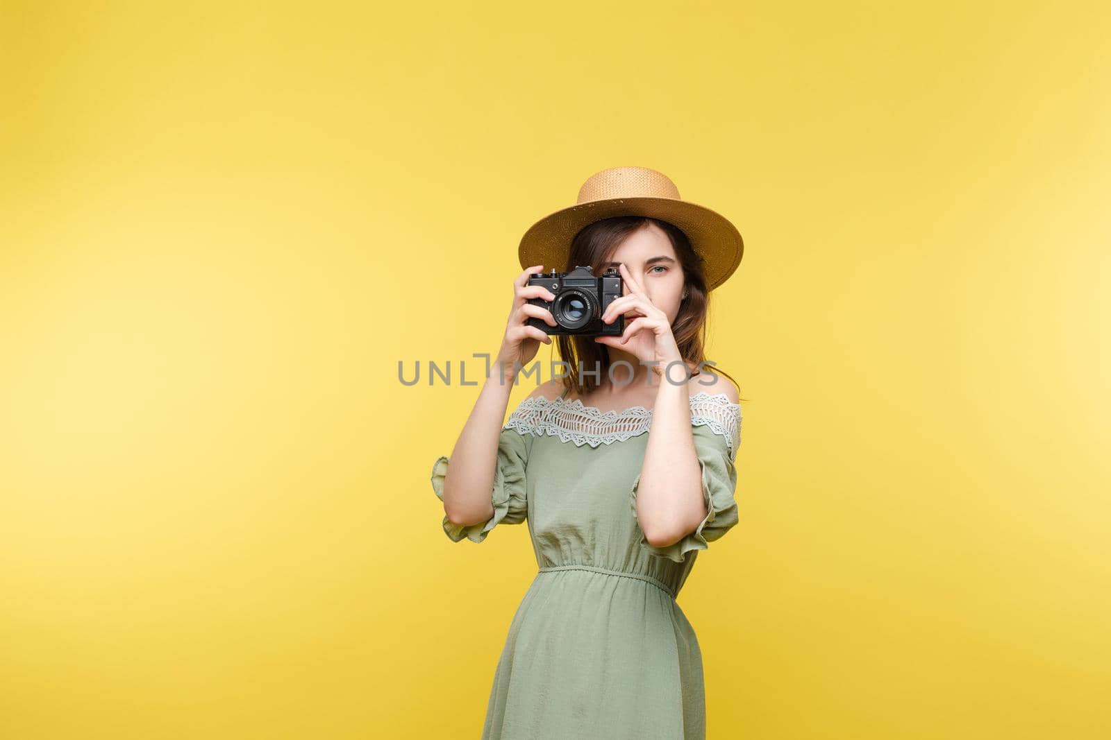 Front view of bright young girl wearing straw hat and glasses posing on yellow isolated background in studio. Female tourist keeping camera, looking at camera and smiling. Concept of summer.