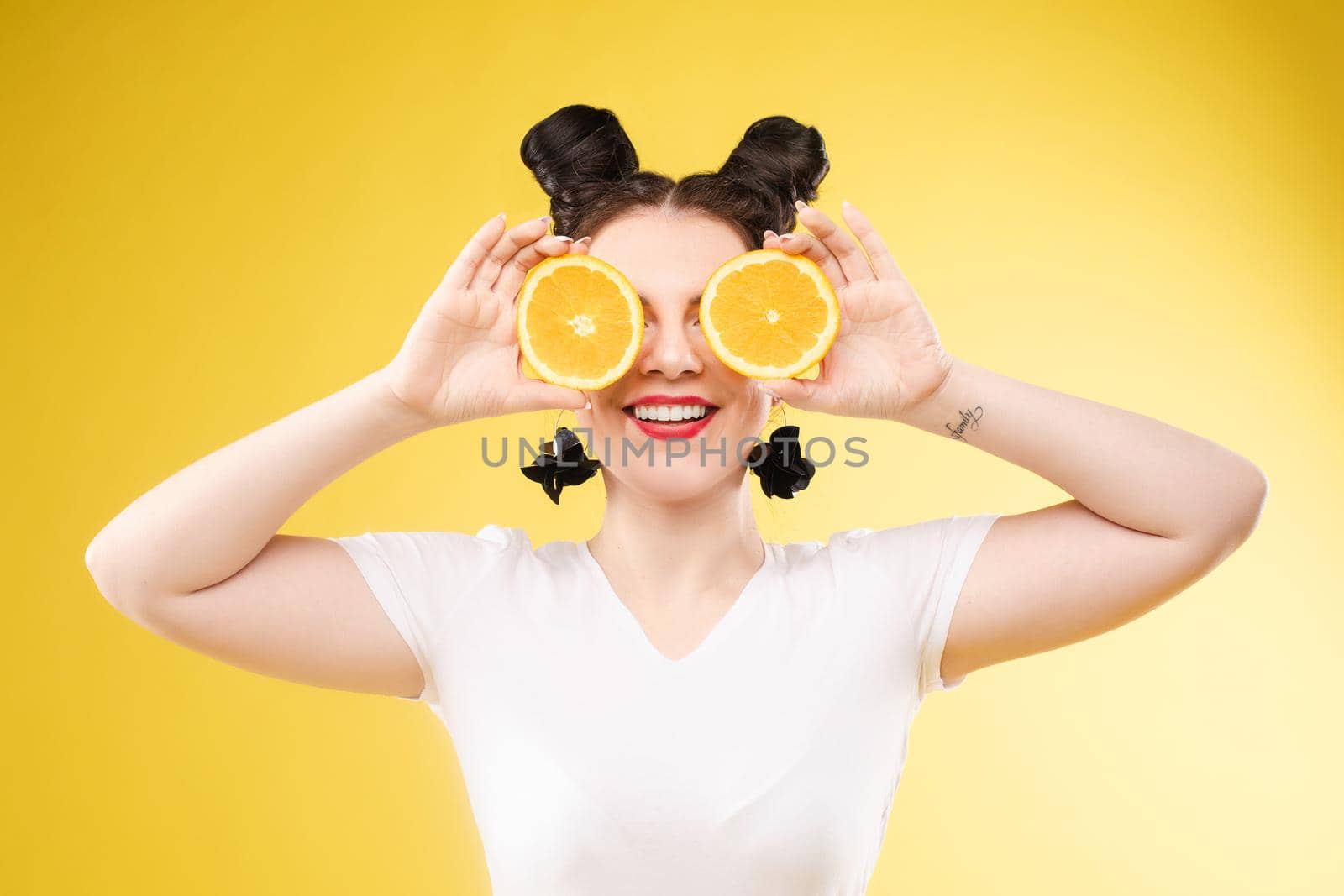 Studio headshot of young funny brunette with hairstyle and red lips holding halved oranges on eyes against bright yellow background.