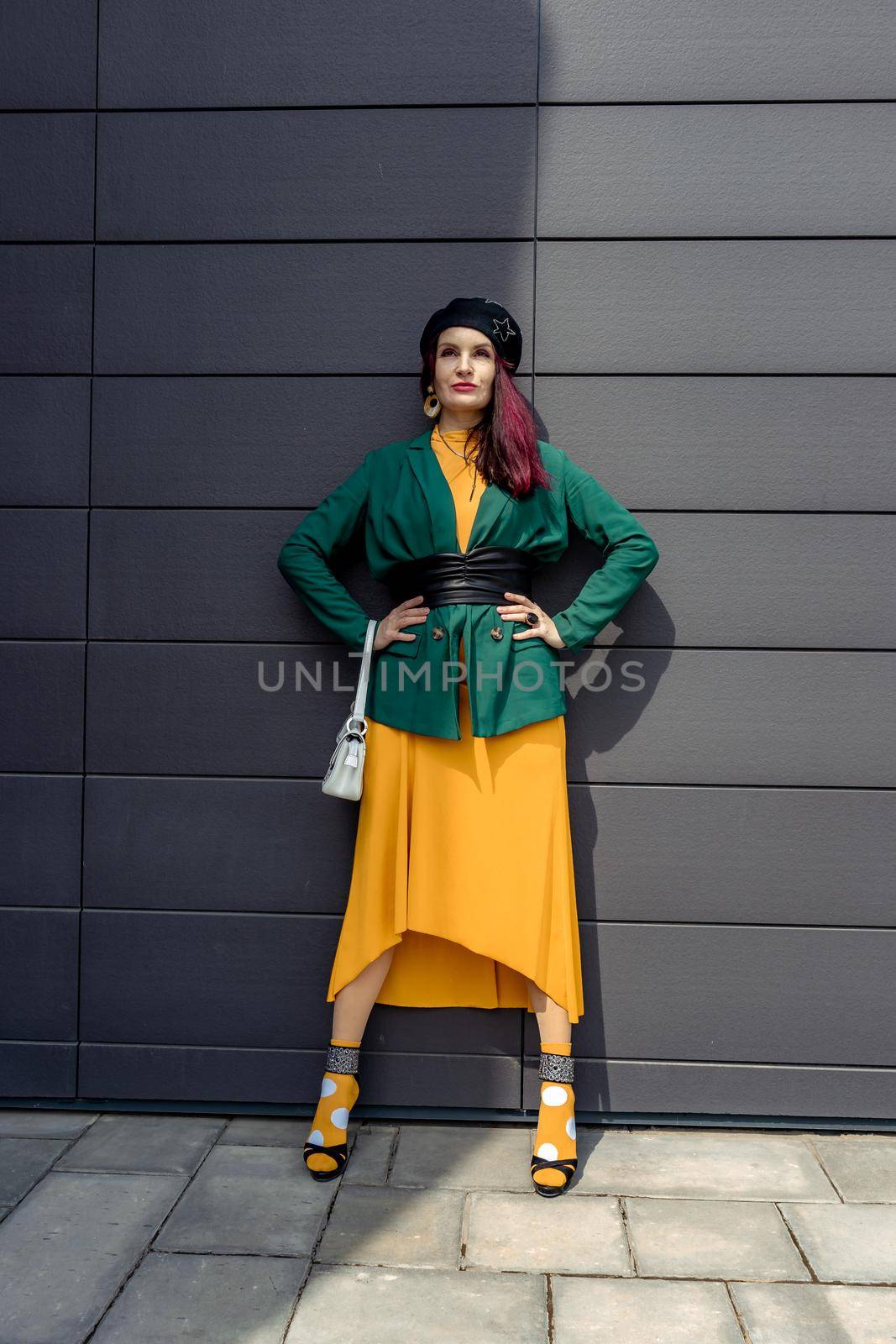 Young woman in bright clothes, yellow skirt and green jacket. Yellow socks in sandals, beret on the head, hair with the color of magenta. Caucasian female fashion model standing against gray wall background, open with copy space. by Matiunina