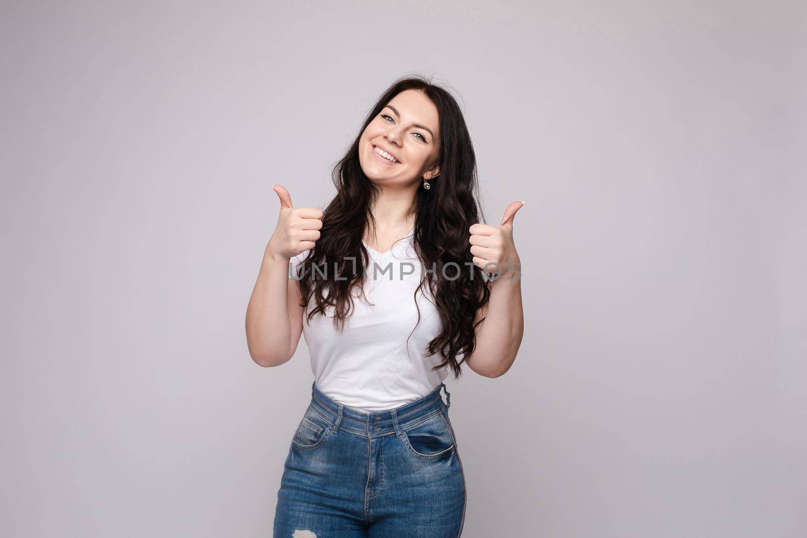 Front view of positive blonde wearing white shirt and jeans looking at camera and showing thumbs up. Young woman posing and laughing on grey isolated background. Concept of happiness.