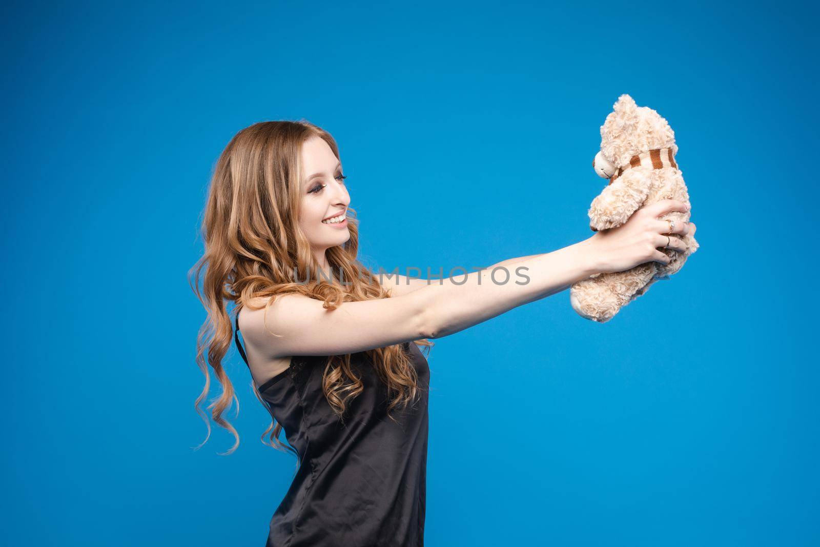 Studio portrait of gorgeous young woman with long wavy hair holding lovely teddy bear in outstretched arms on deep blue background.