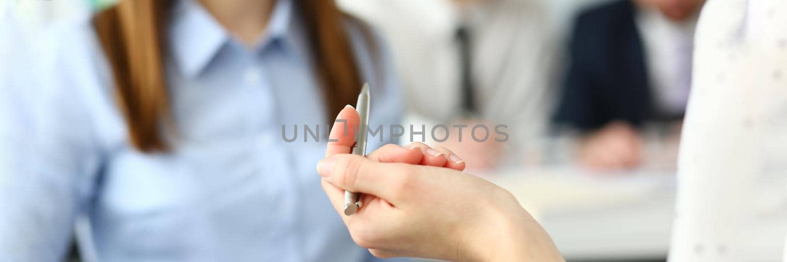 Focus on beautiful woman arm holding writing pen. Pretty female in classy blouse talking with friendly colleagues about important business project. Company meeting concept
