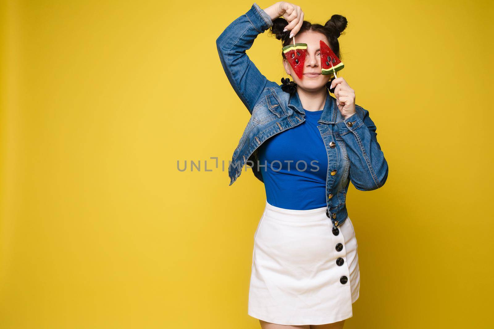 Full length studio portrait of laughing model in white top and green skirt and heels holding two sweet candies on her eyes like sunglasses with her mouth open. Isolate on yellow.