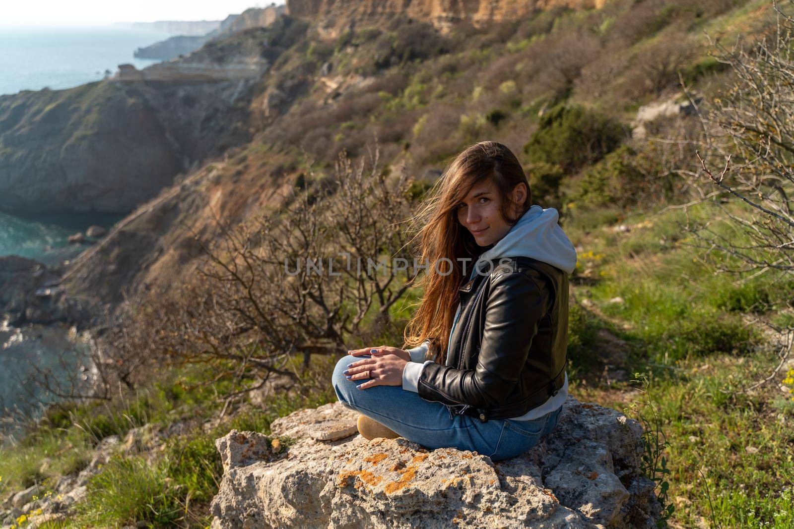 Woman tourist enjoying the sunset over the sea mountain landscape. Sits outdoors on a rock above the sea. She is wearing jeans, a blue hoodie and a black leather jacket. Healthy lifestyle, harmony and meditation by Matiunina