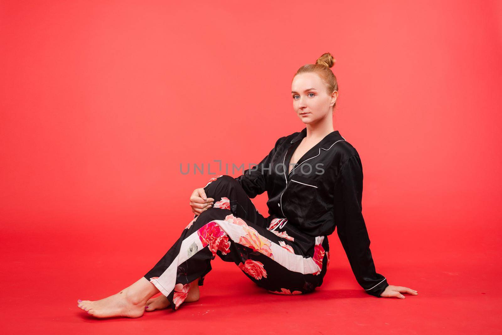 Full length portrait of attractive woman with red hair in a bun wearing a silk black pyjamas with floral trousers. She is sitting on the floor over red background and looking away.