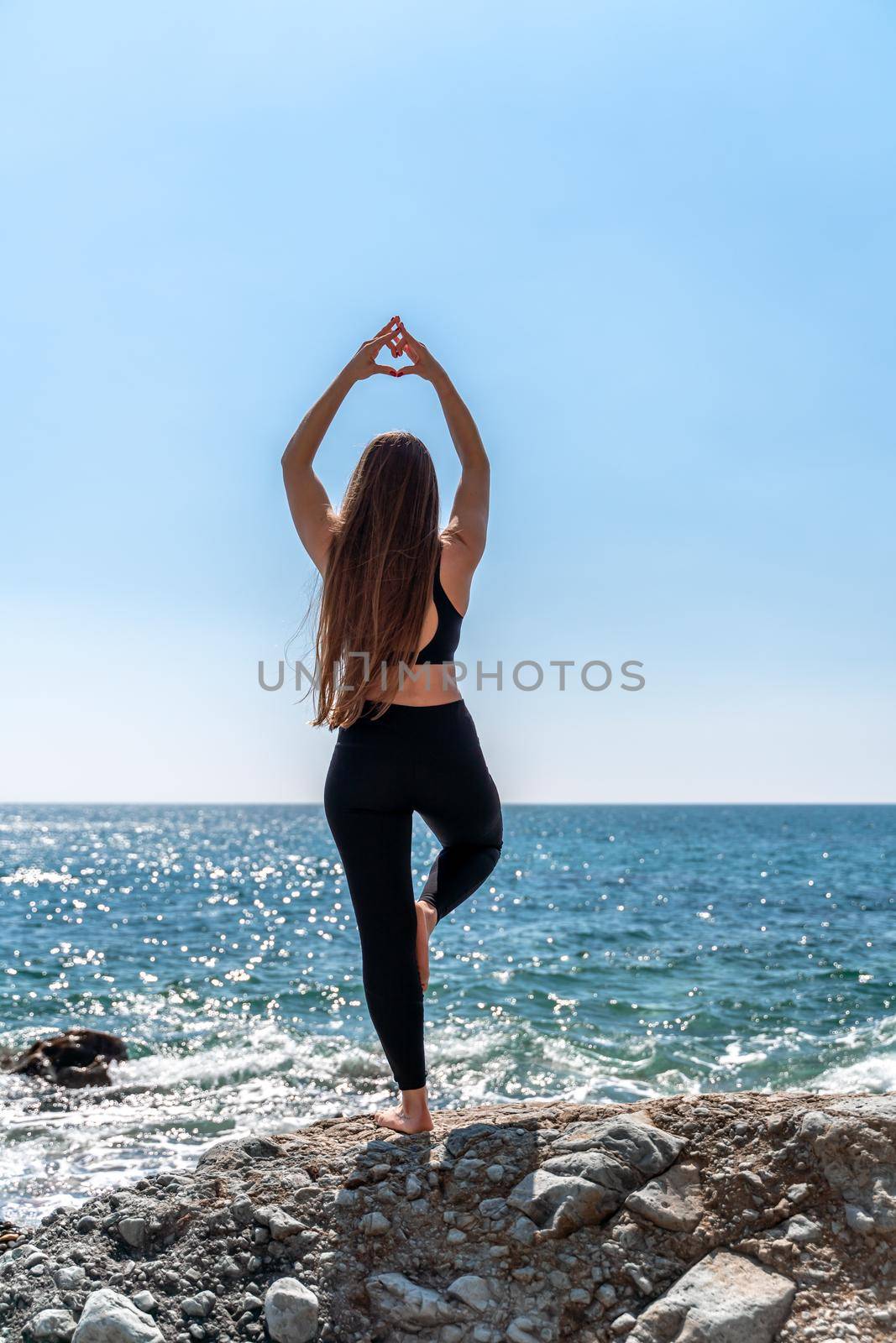 Young woman in black leggings and black top with long flowing hair practicing stretching outdoors by the sea on a sunny day. Women's yoga, fitness, pilates. The concept of a healthy lifestyle, harmony. by Matiunina