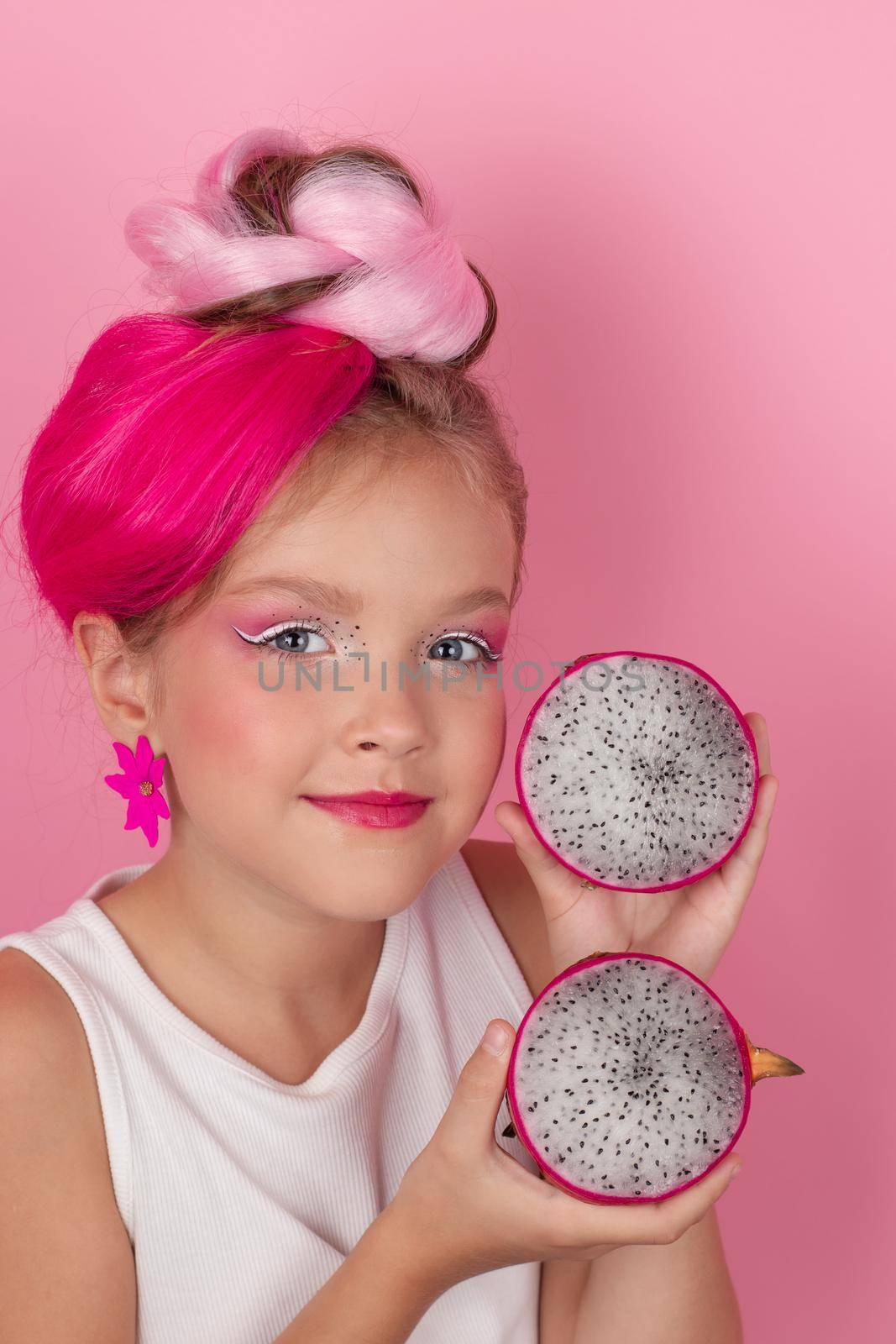 Close-up portrait of pretty girl with pink hairstyle with dragon fruit on pink background. Studio shot of charming tween girl with pink make up enjoying juicy red pitaya. exotic Pitahaya fruit.