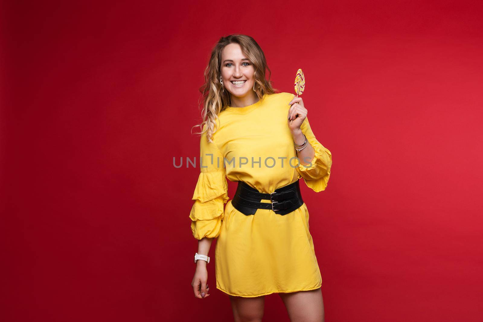 Studio portrait of attractive young woman with long wave hair enjoying tasty sweet colorful lollipop with her eyes closed. She is wearing bright yellow blouse and fashionable earrings. Isolate on red.