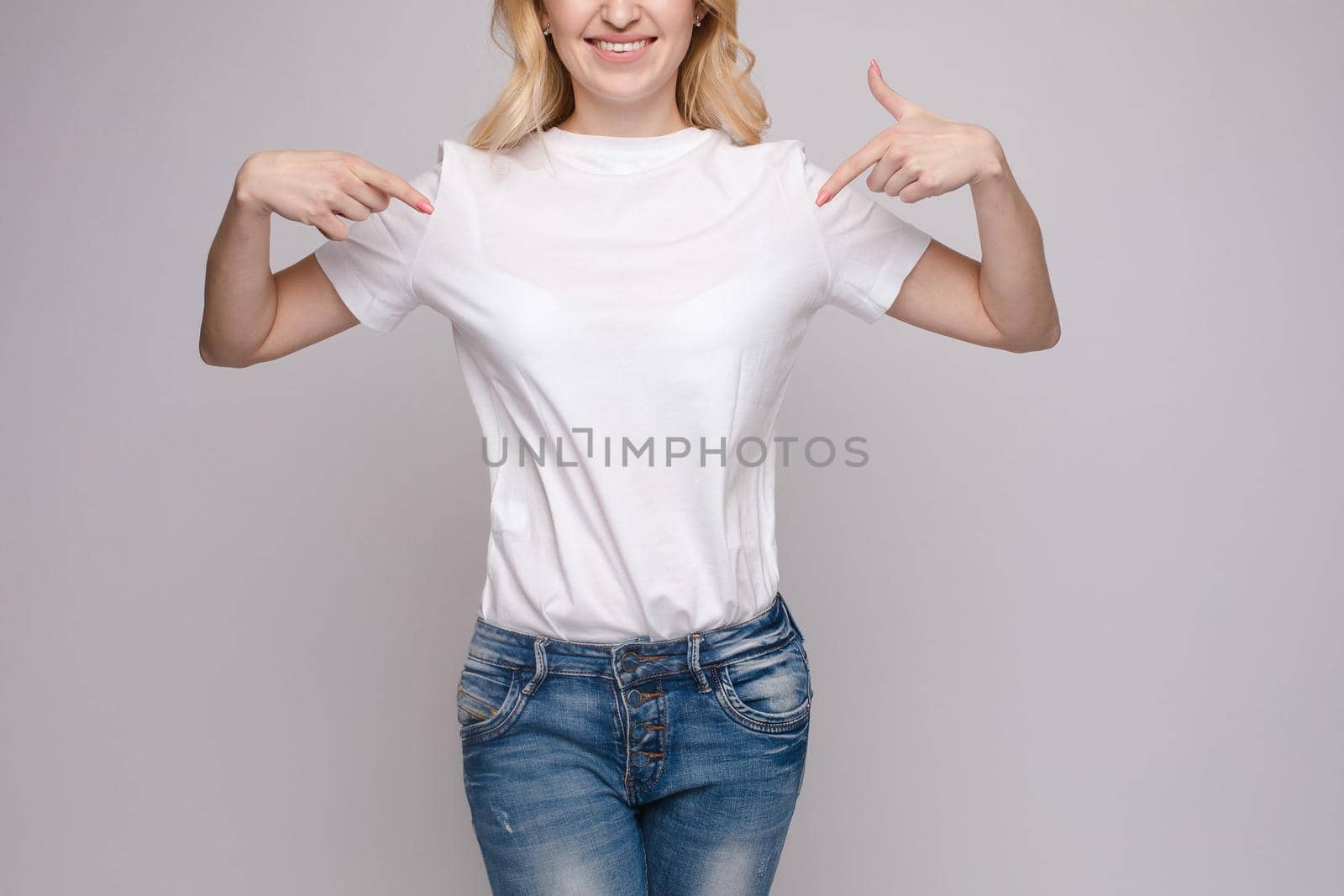 Cropped view from front of happy brunette standing in white shirt and jeans and pointing at outfit with fingers. Cheerful girl laughing and posing on grey isolated backround. Concept of casual style.