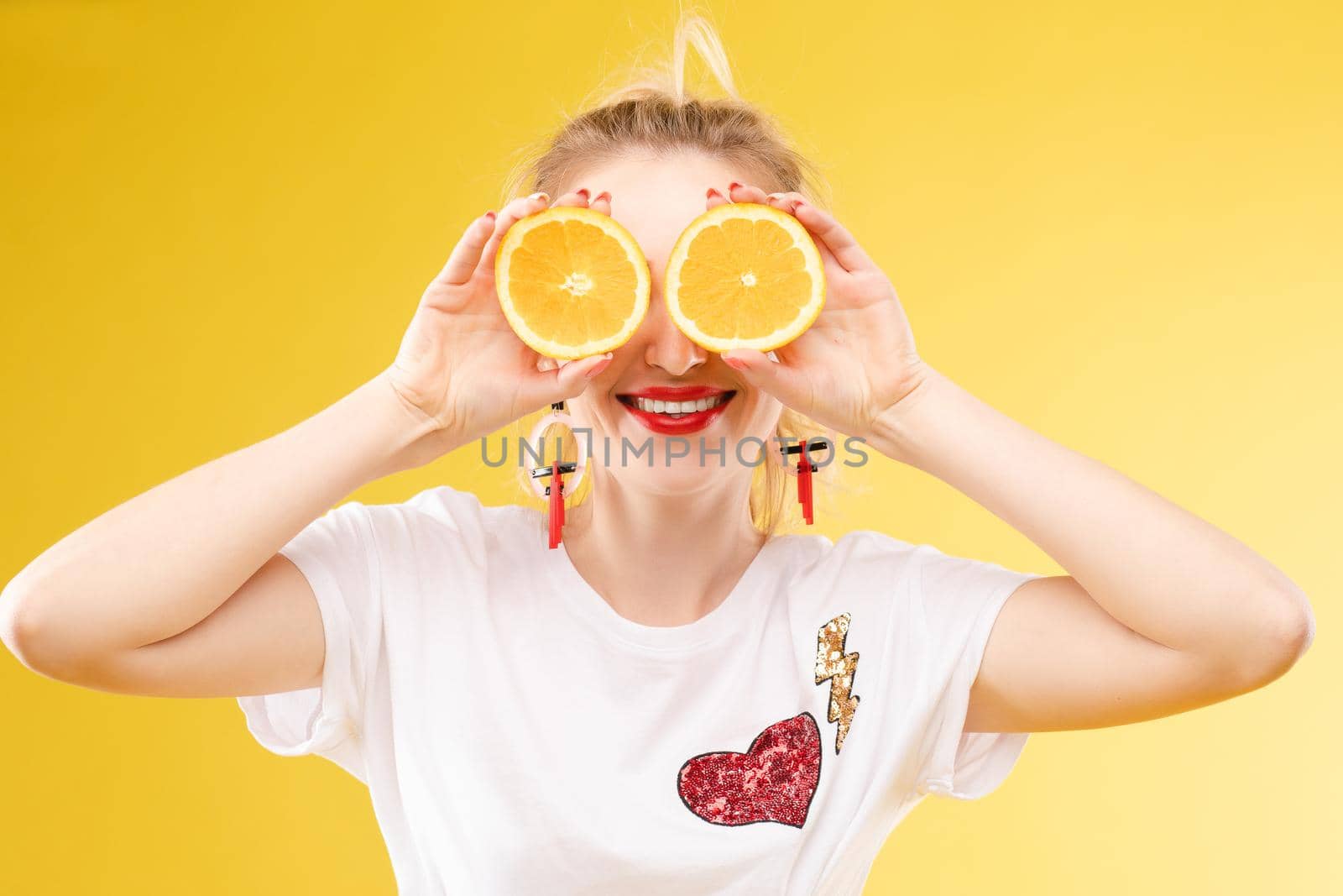 Front view of cheerful young blonde posing with fresh oranges on isolated background. Funny girl keeping fruit and closing eyes in studio. Concept of happiness and health.