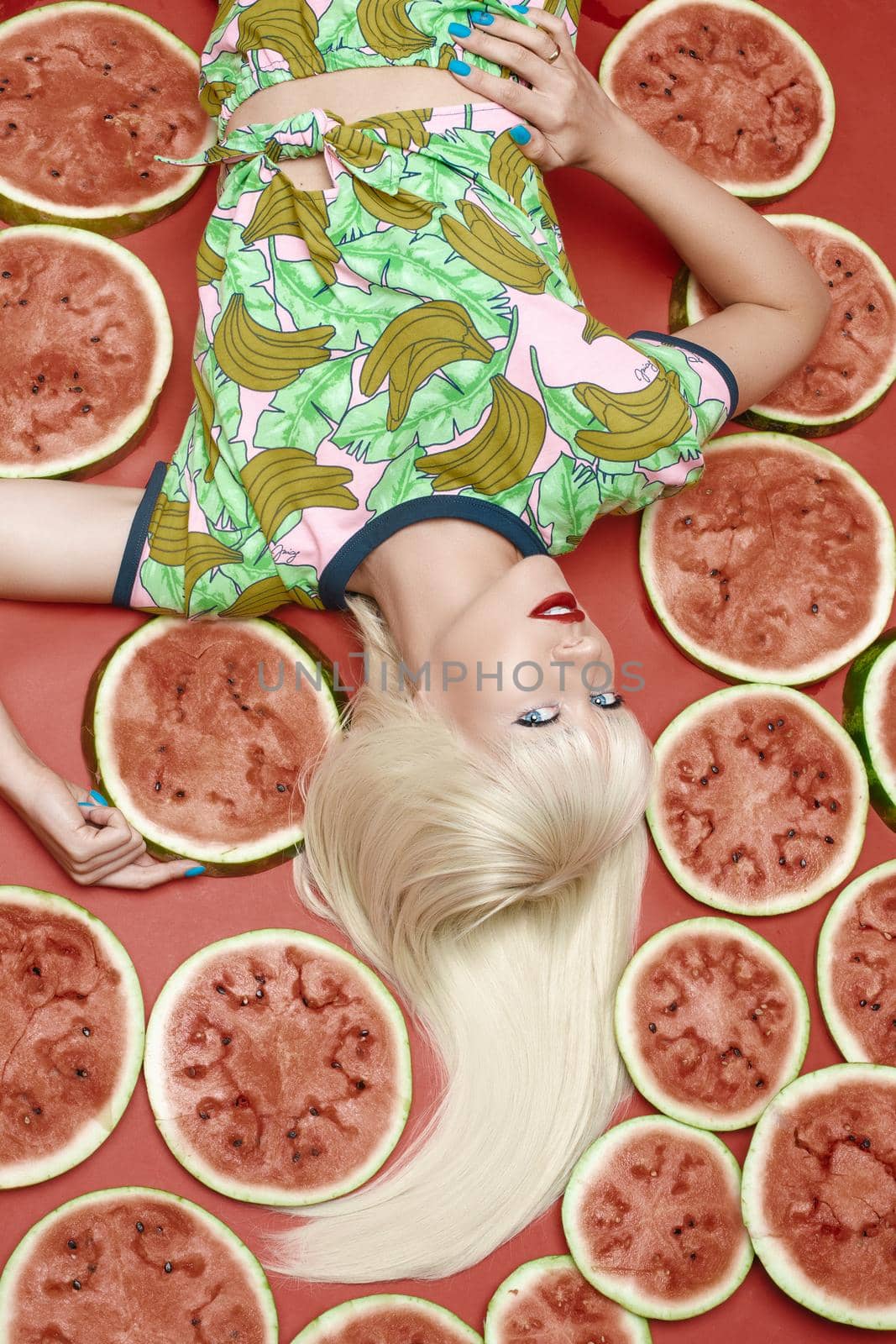 View from above fashionable blonde with makeup lying near fresh tasty watermelon. Attractive female model looking at camera and posing with sweet fruits. Concept of summer and brightness.