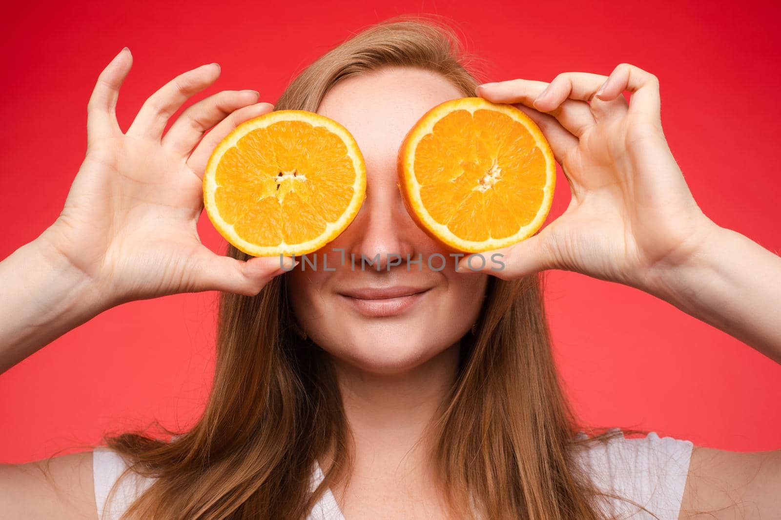 Studio headshot of young funny brunette with hairstyle and red lips holding halved oranges on eyes against bright yellow background.