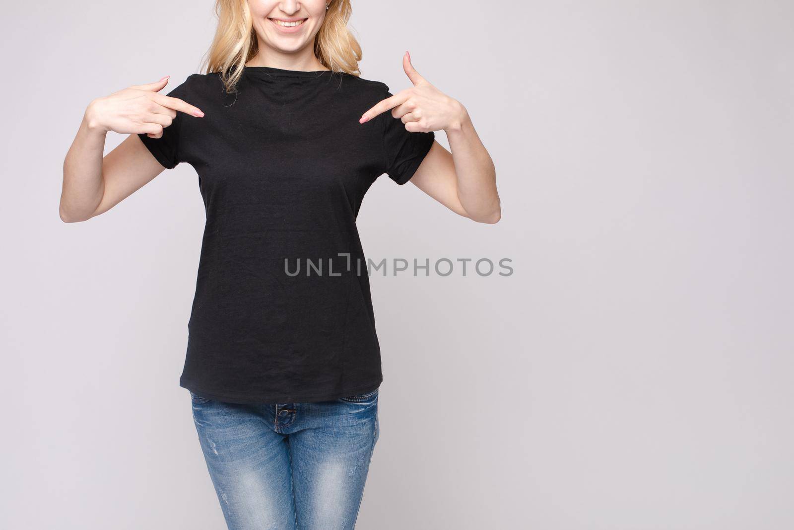 Cropped view from front of happy blond standing in black shirt and jeans and pointing at outfit with fingers. Cheerful girl laughing and posing on grey isolated backround. Concept of casual style.