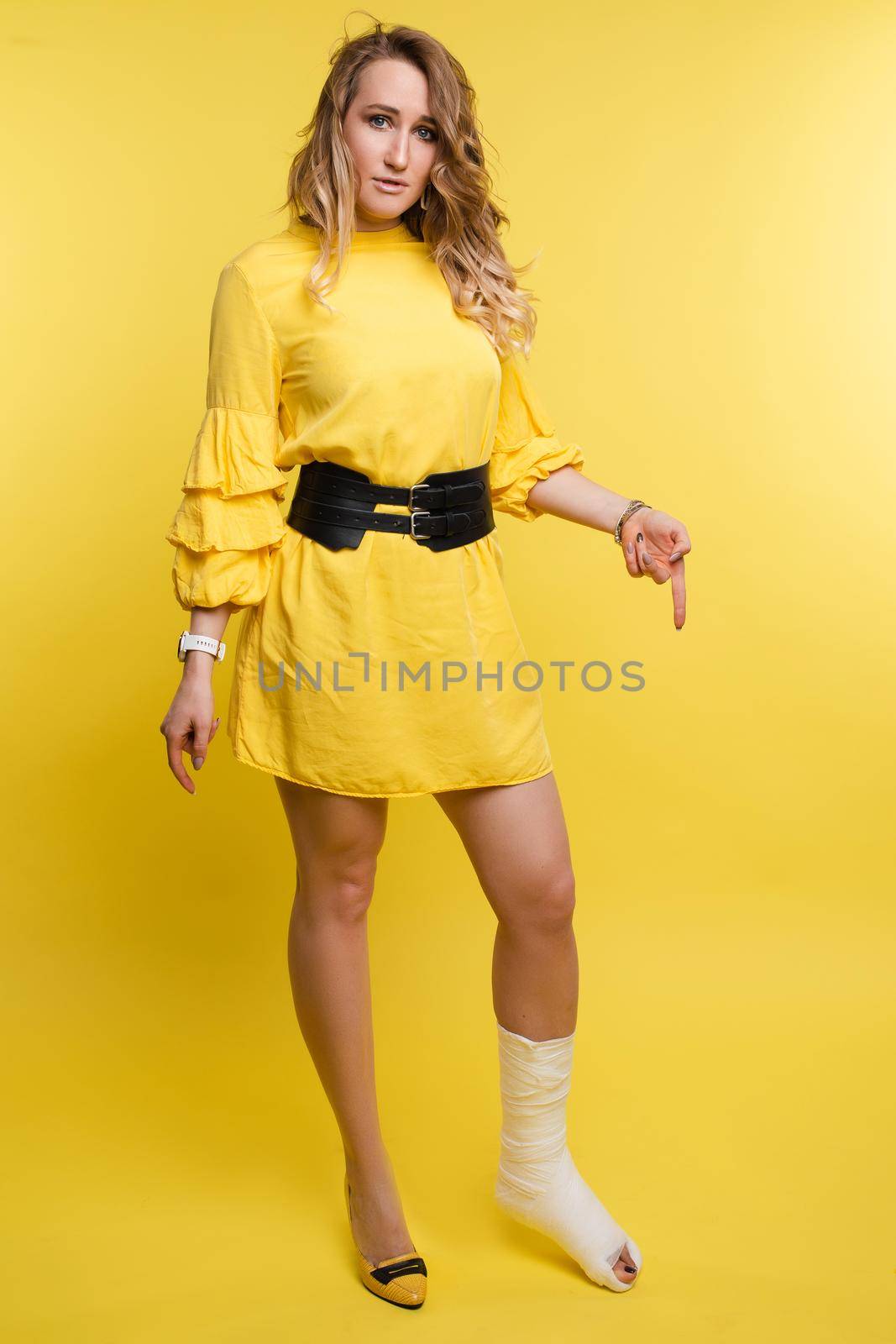 Full length studio portrait of beautiful caucasian woman in yellow dress with black belt pointing at her broken leg in plaster. Isolate on yellow background. Injury concept.