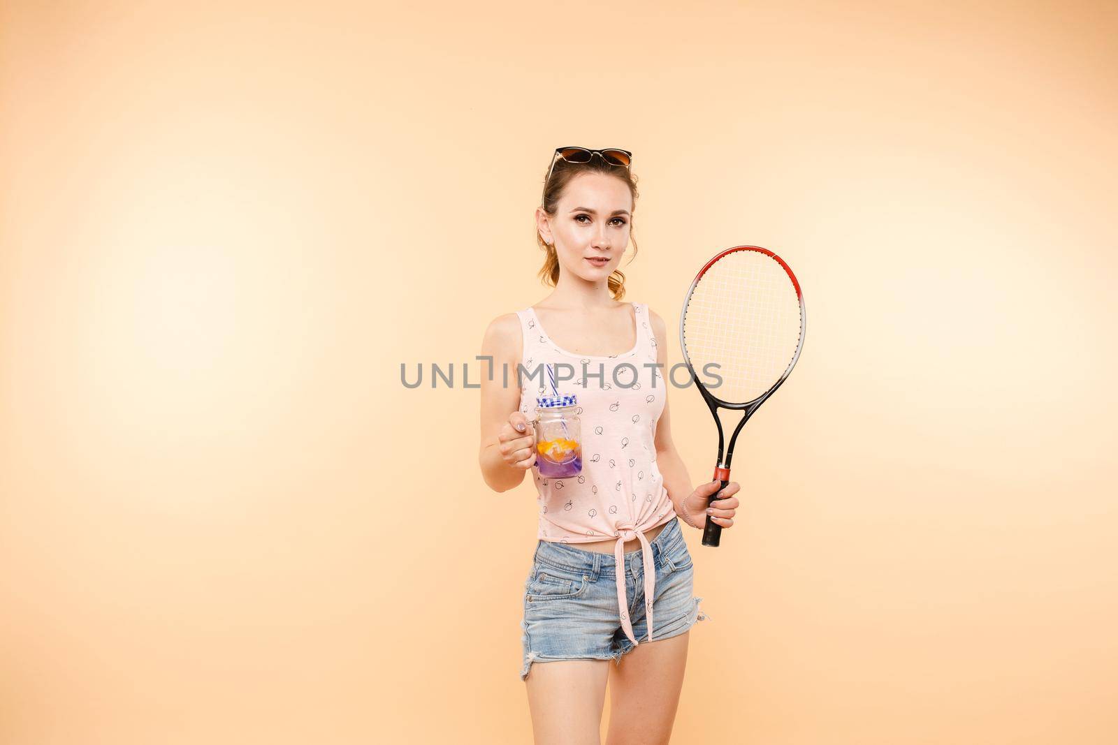 Active young girl in t-shirt and shorts holding racket in hand. Brunette lady with sunglasses on head enjoying drink after playing tennis. Beautiful woman relaxing and drinking lemonade after game.