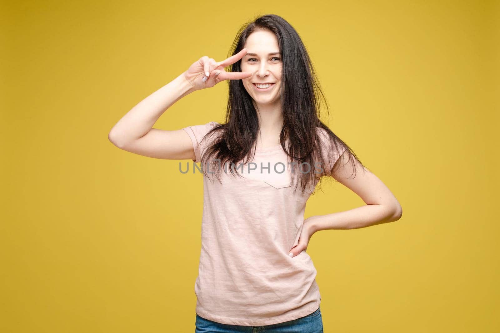 Studio portrait of charming brunette young woman in white top and green skirt holding peace sign near her eye. She is smiling at camera. Isolate on orange.