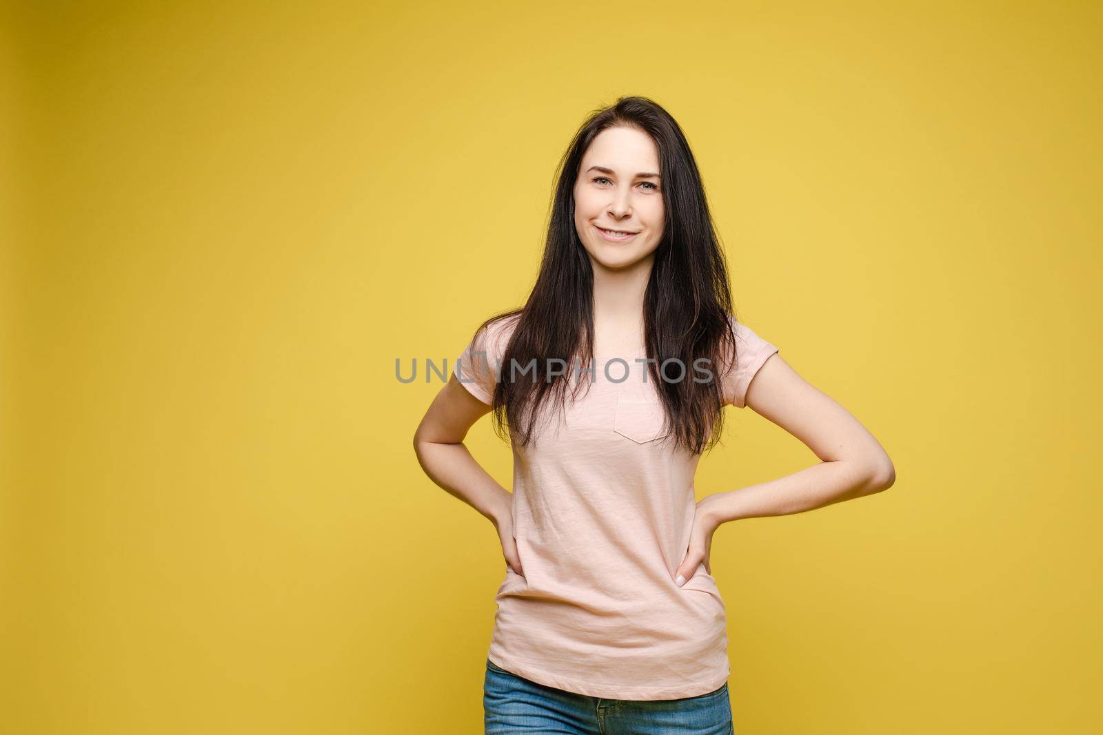 View from front of beautiful slim woman wearing white shirt and jeans standing steady on frey isolated background. Young blonde looking at camera, smiling and posing in studio.