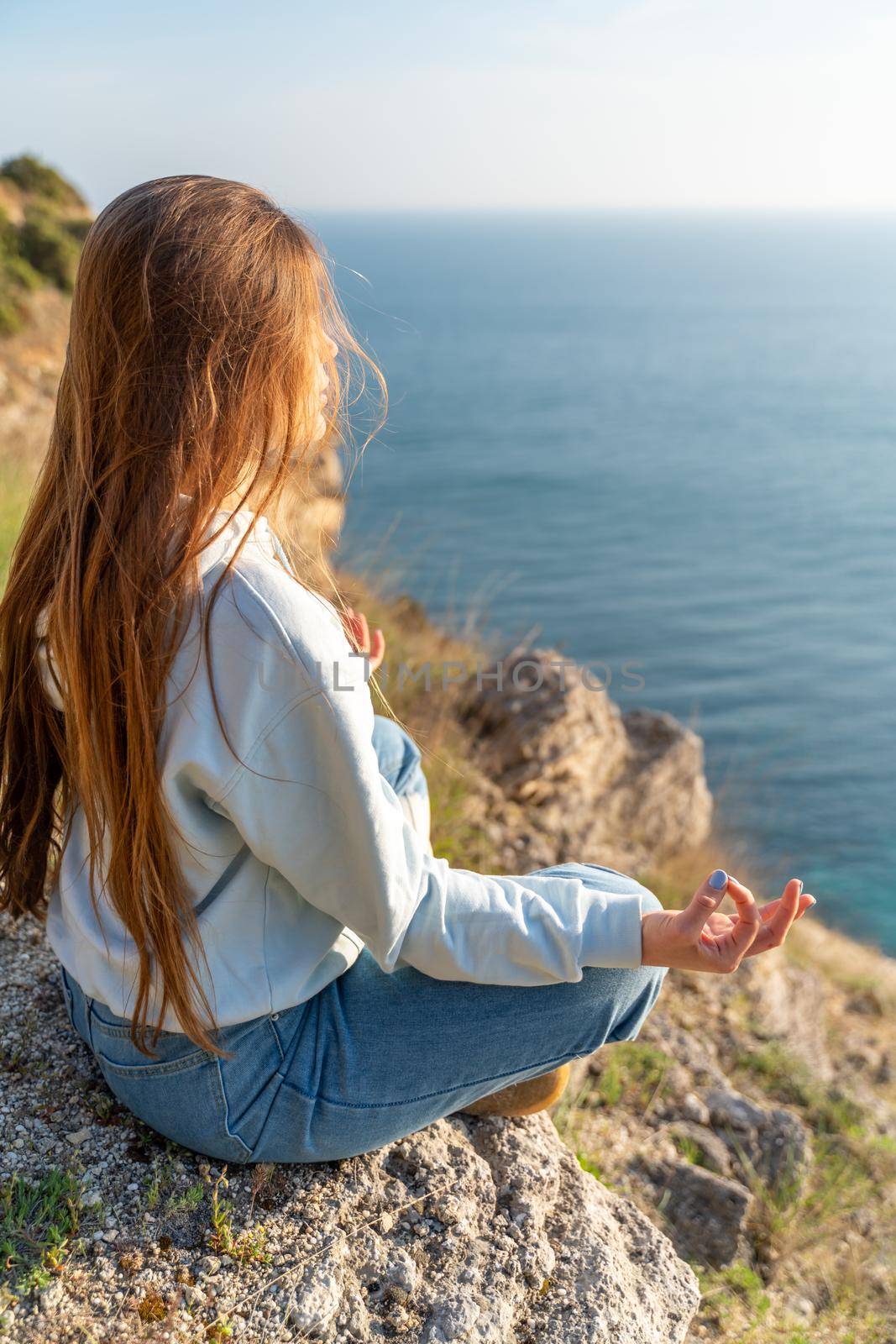 Woman tourist enjoying the sunset over the sea mountain landscape. Sits outdoors on a rock above the sea. She is wearing jeans and a blue hoodie. Healthy lifestyle, harmony and meditation.