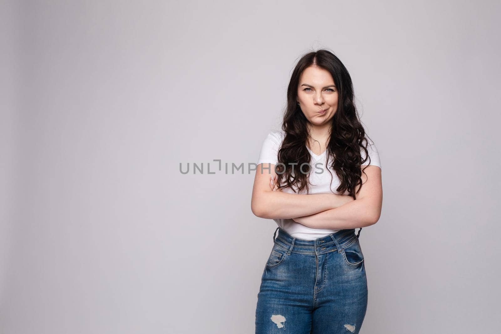 Studio portrait of unsatisfied young brunette caucasian woman with wavy hair in overall with colorful pattern holding arms folded and looking at camera with grief, dissatisfaction, anger and disbelief. Isolate.