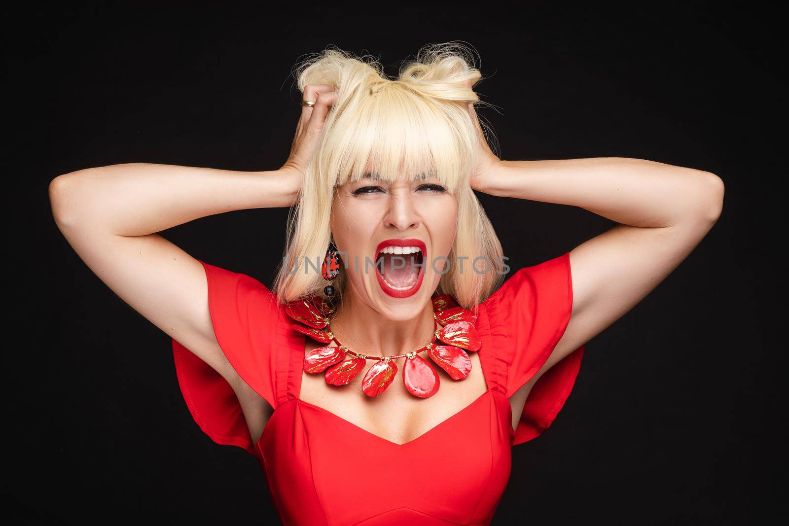 Fashion stressed blonde woman in red dress shout with open mouth holding head by hand medium closeup. Attractive worried female model posing having negative emotion isolated at black studio background