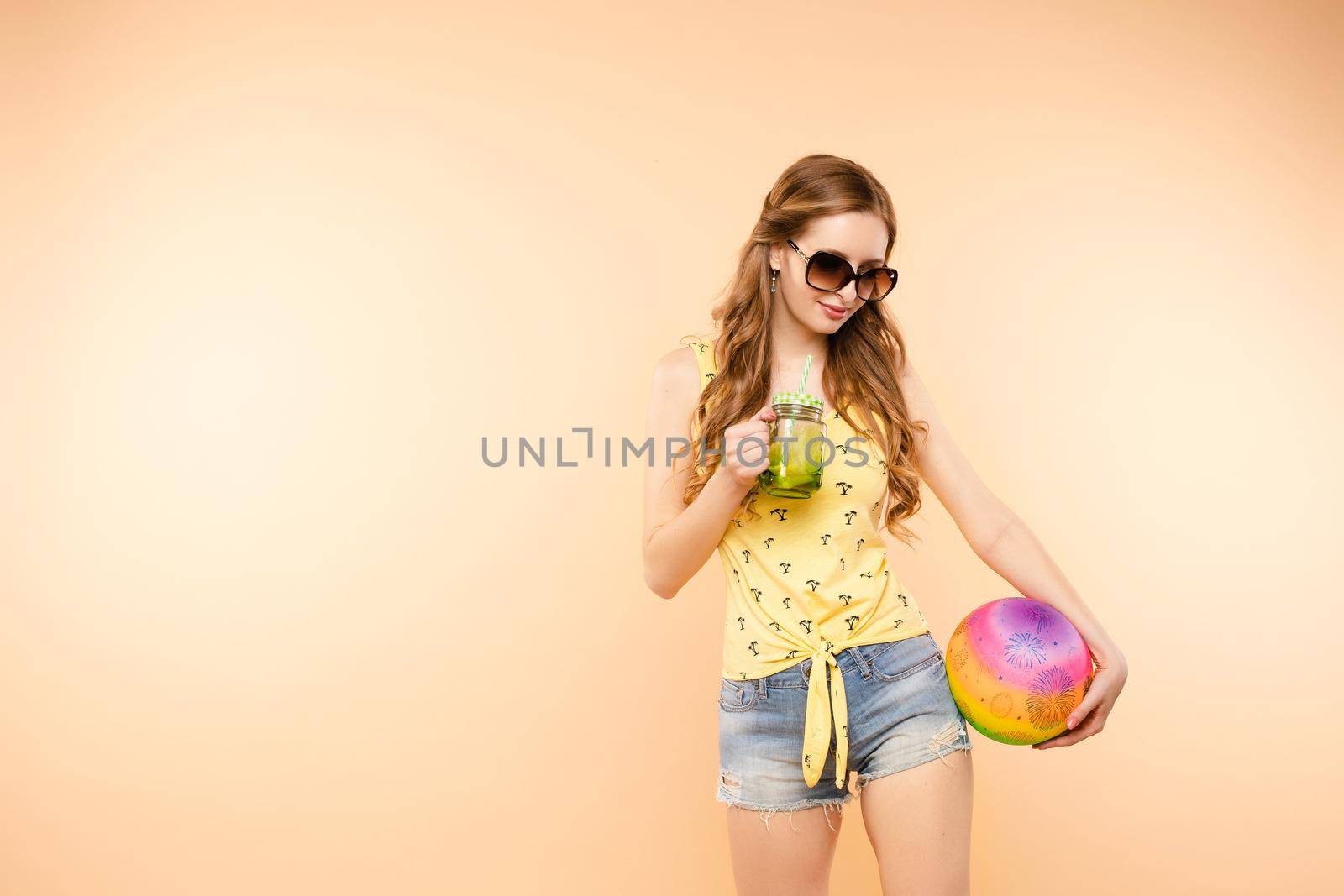 Side view of sporty blonde drinking fresh juice, doing sport and posing in studio. Young model keeping ball and racket and smiling on isolated background. Concept of energy and power.