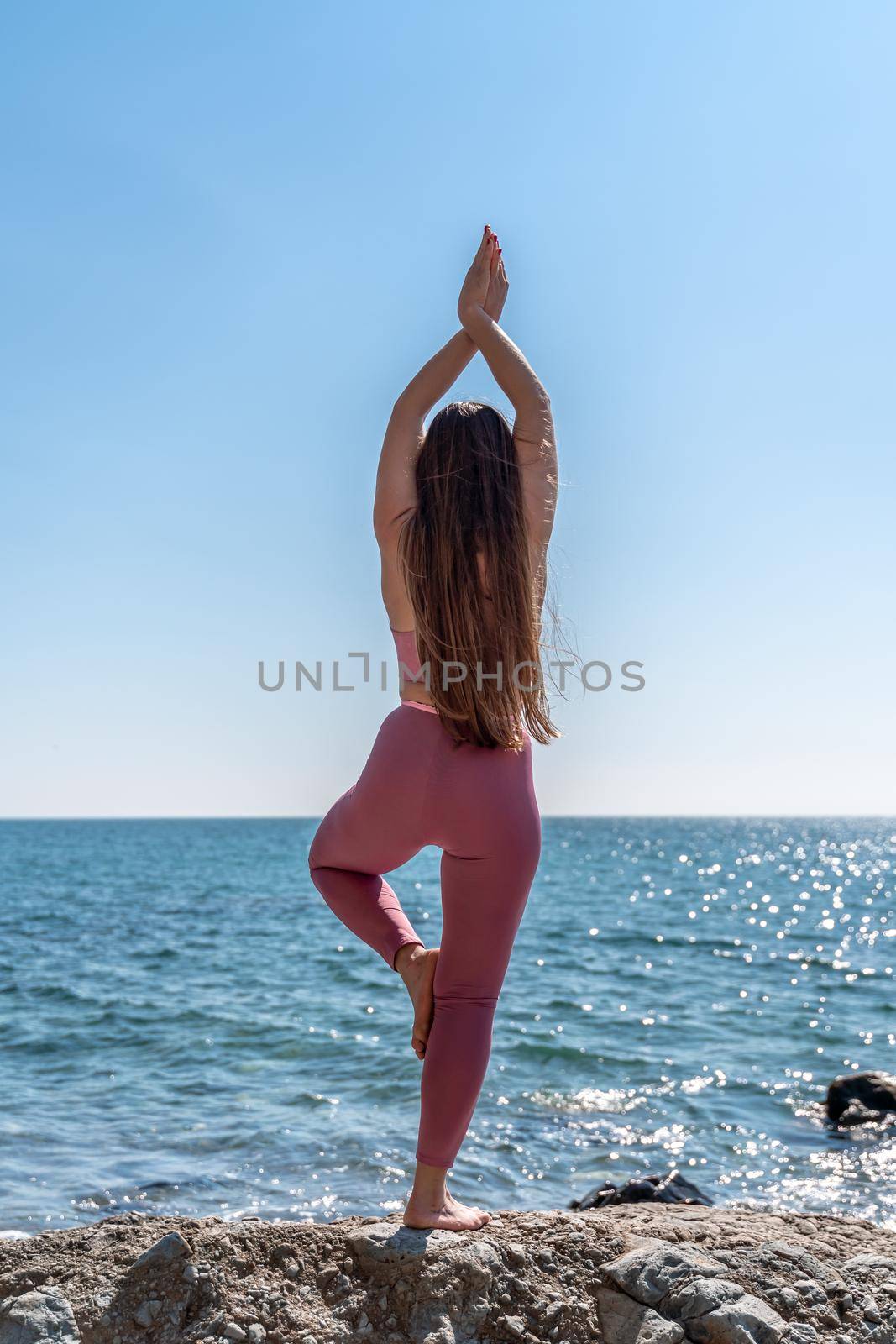 A young woman in pink leggings and a pink top with long loose hair practices stretching outdoors by the sea on a sunny day. Women's yoga, fitness, Pilates. The concept of a healthy lifestyle, harmony