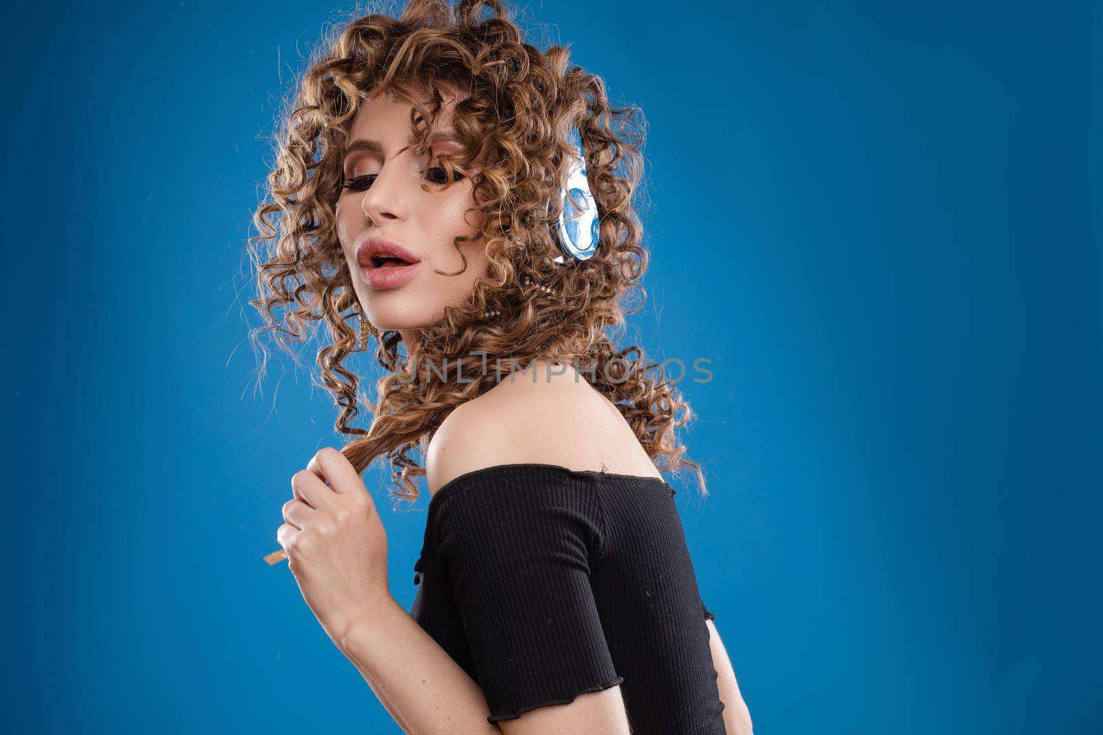 Front view of crazy curly woman with makeup wearing headphones and listening music on isolated background. Pretty girl dancing, singing and enjoying song in studio. Concept of disco and party.