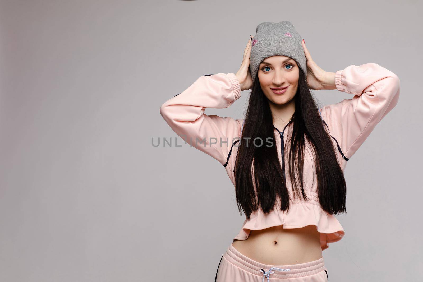 Beautiful girl with big blue eyes and opened belly posing at camera. Brunette happy girl with long hair in pink costume smiling. Stylish model in sportswear and grey hat holding head by both hands.