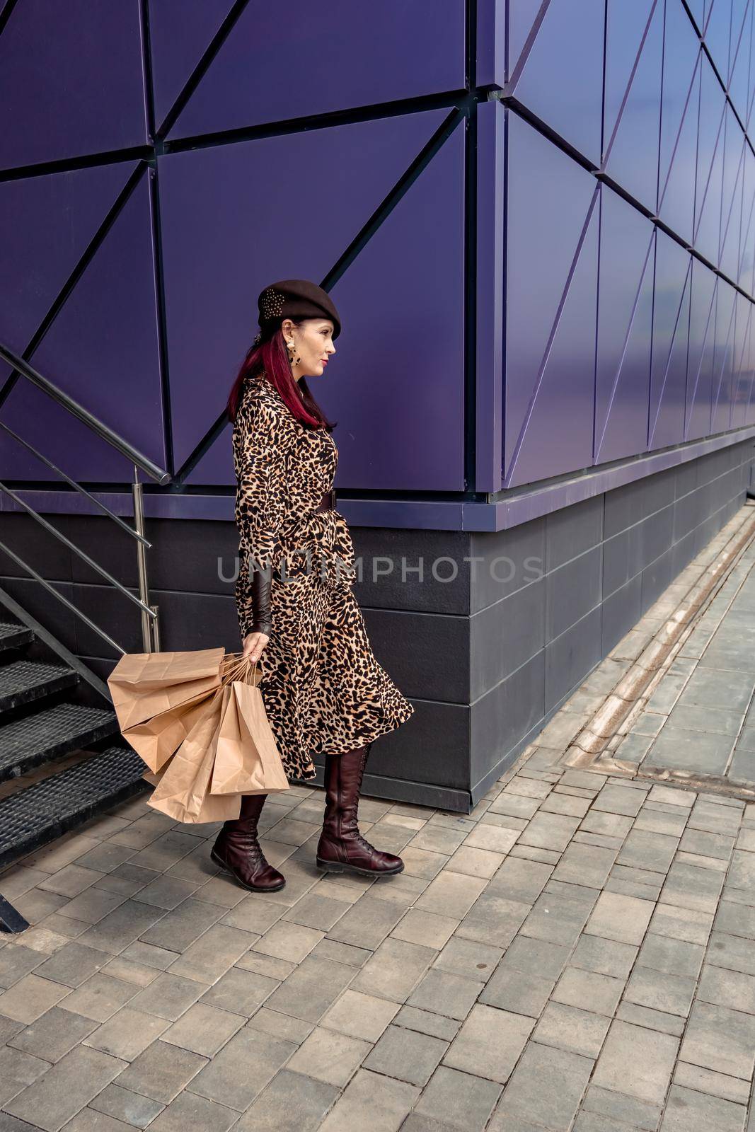 A happy shopaholic girl keeps her bags near the shopping center. A woman near the store is happy with her purchases, holding bags. Dressed in a leopard print dress. Consumer concept. by Matiunina