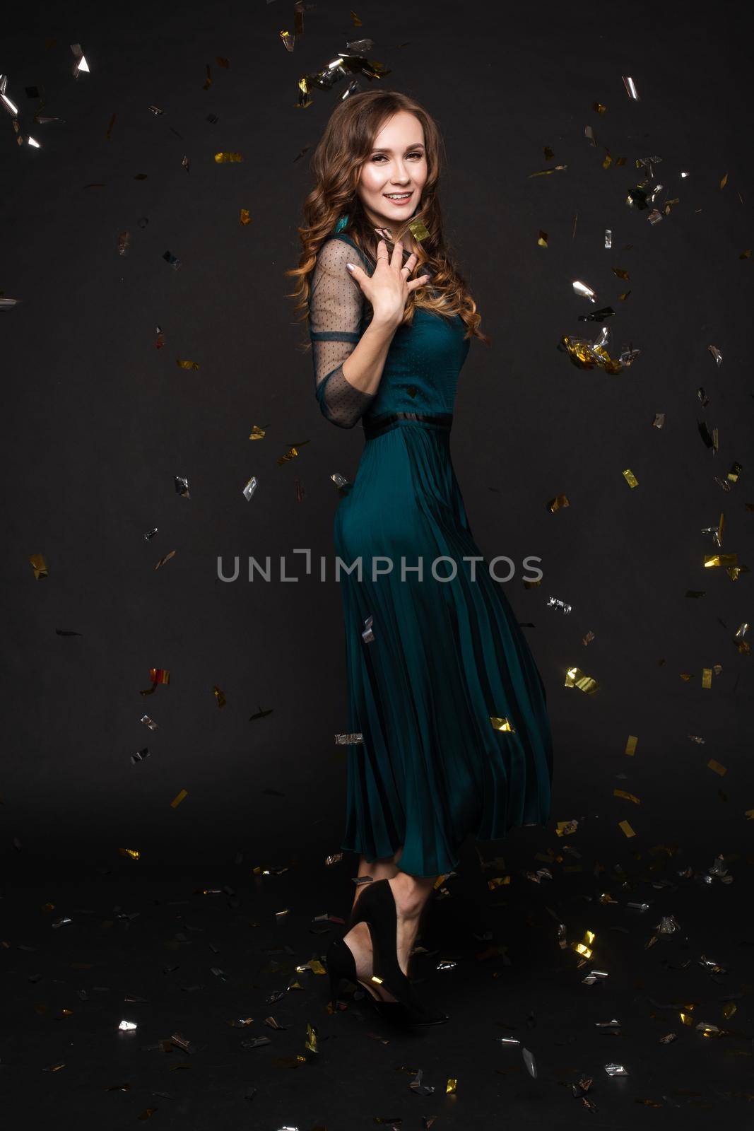 Happy young girl looking surprisingly while golden confetti falling. Beautiful woman in emerald elegant long dress smiling and dancing among decorations. Gorgeous lady celebrating birthday on party.