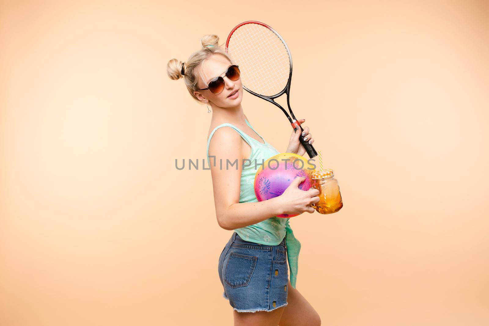 Side view of sporty blonde drinking fresh juice, doing sport and posing in studio. Young model keeping ball and racket and smiling on isolated background. Concept of energy and power.