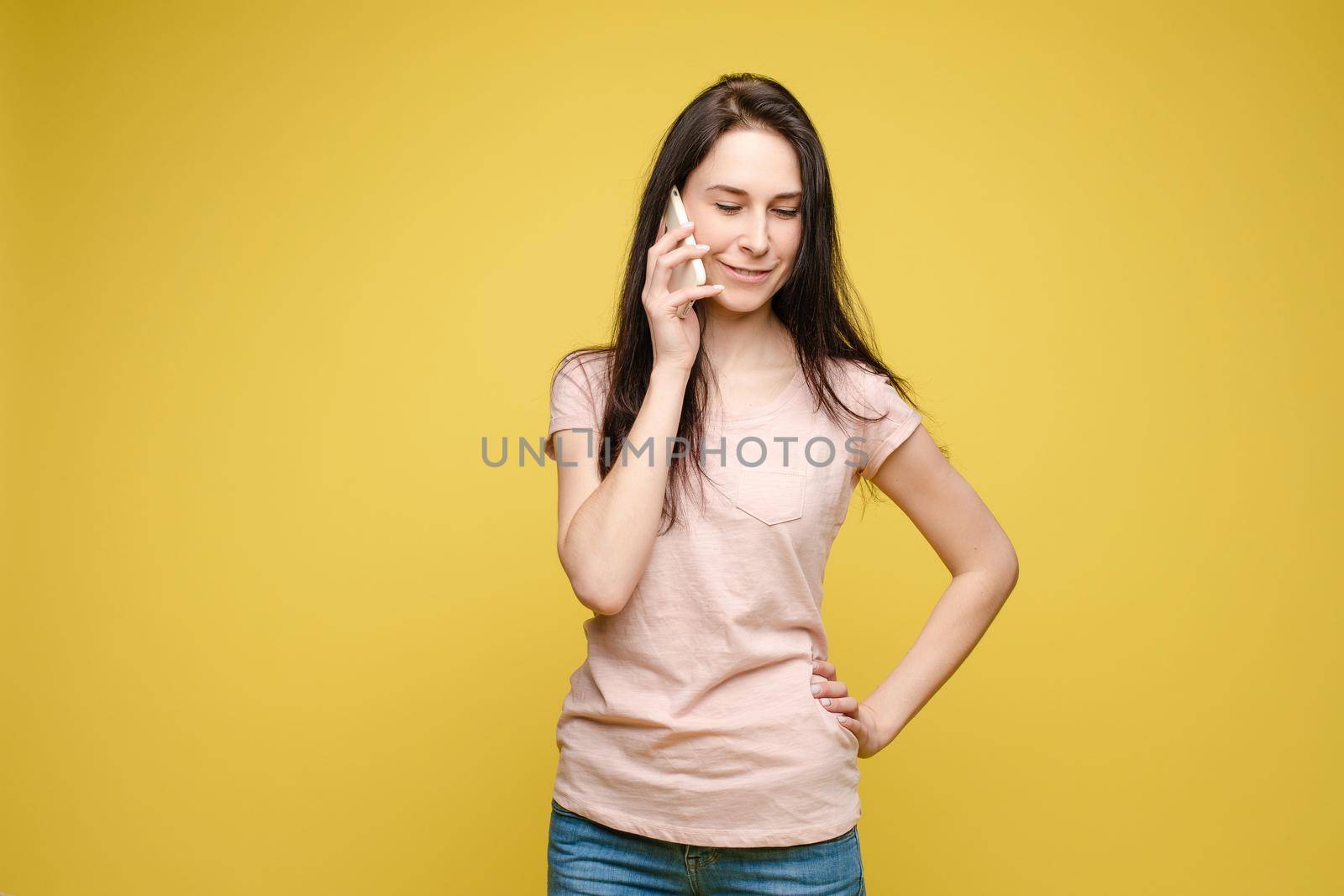 Front view of stylish young girl in bright light dress talking by phone on yellow isolated background in studio. Lovely female chattering and smiling. Concept of communication and happiness.