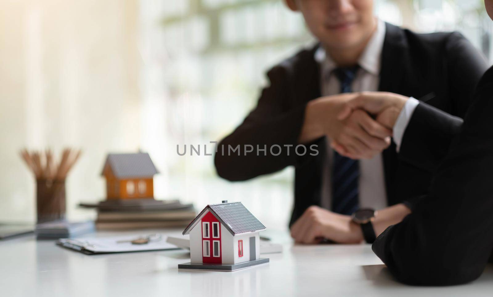 Estate agent shaking hands with his customer after contract signature, Contract document and house model on wooden desk.