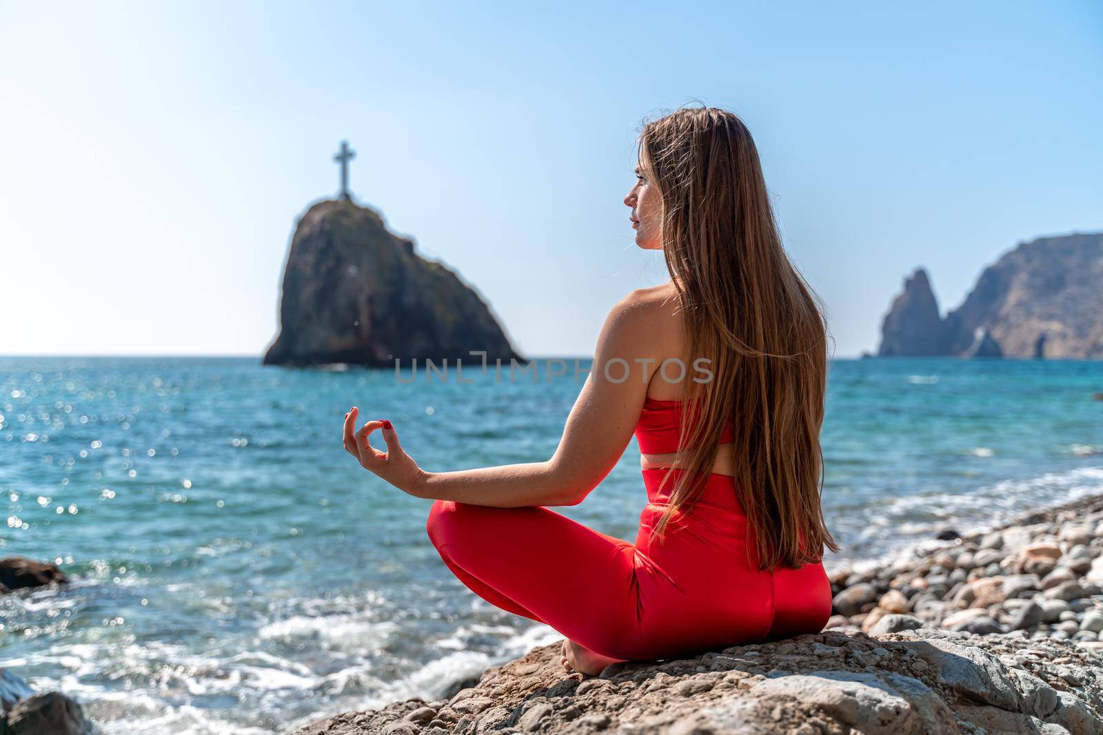 A young woman in red leggings and a red top with long loose hair practices yoga outdoors by the sea on a sunny day. Women's yoga, fitness, Pilates. The concept of a healthy lifestyle, harmony