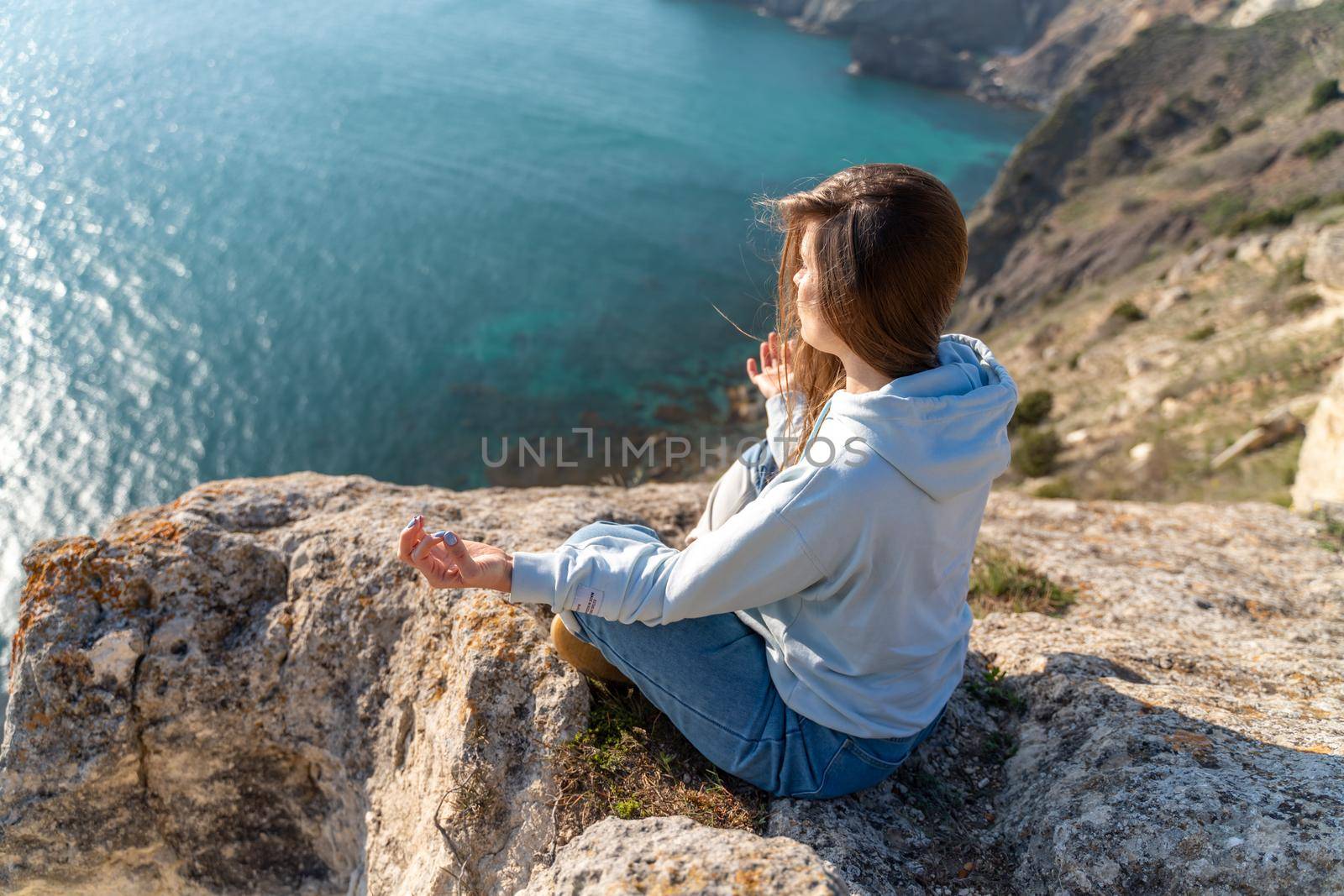 Woman tourist enjoying the sunset over the sea mountain landscape. Sits outdoors on a rock above the sea. She is wearing jeans and a blue hoodie. Healthy lifestyle, harmony and meditation.