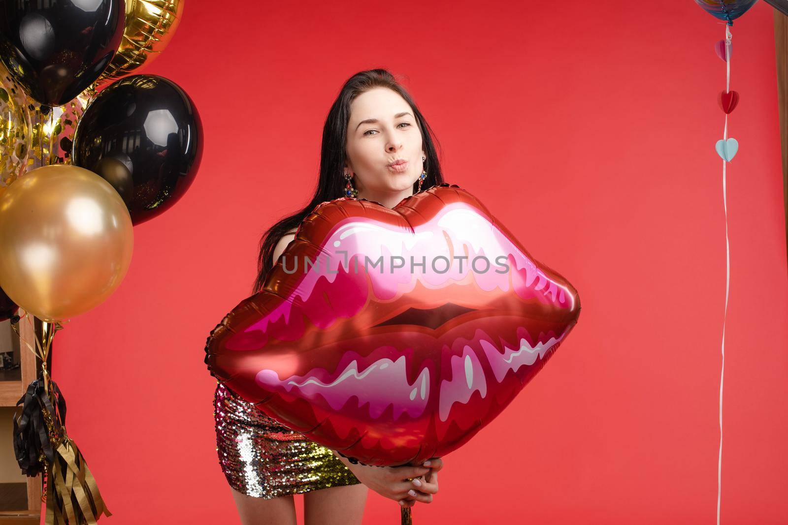 Front view of cheerful attractive girl in bright short dress dancing and laughing on red isolated background. Happy brunette posing near balloons in studio. Smiling woman celebrating new year party.