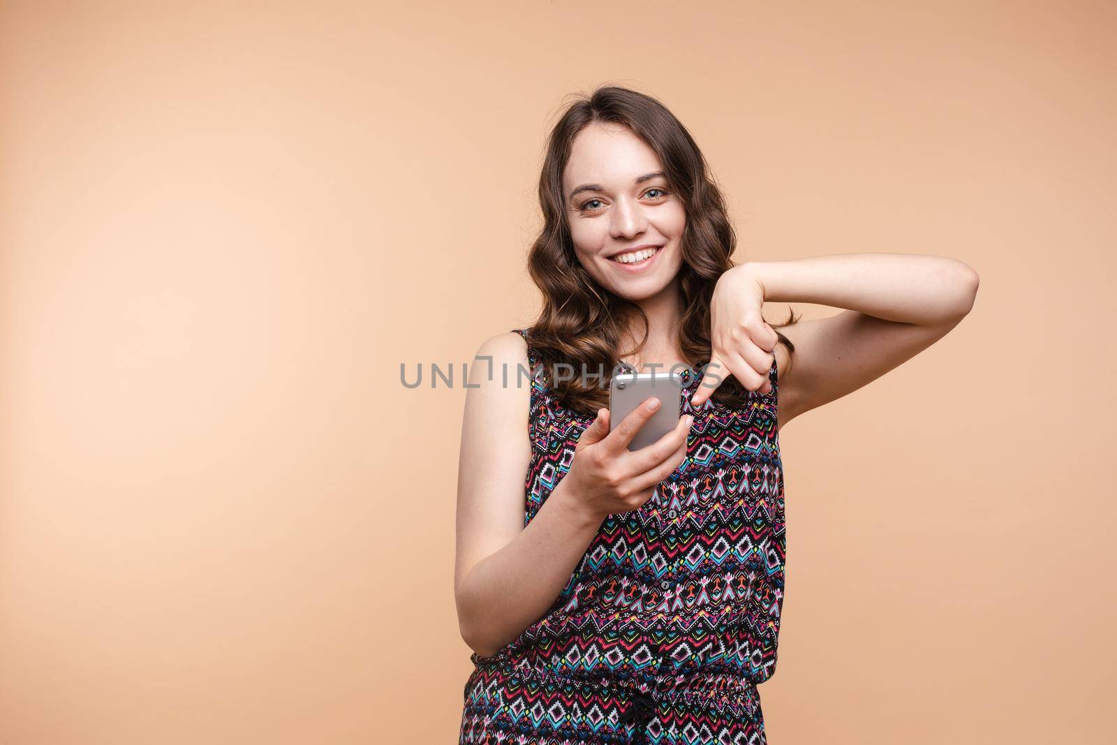 Studio portrait of beautiful caucasian brunette woman in patterned overall pointing at her smartphone with index finger. She is certain or sure about something. I know gesture.